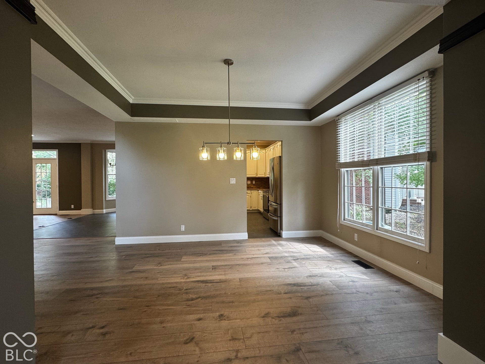 Tray ceiling with crown molding, wood-finish flooring, two large windows, built-in lighting fixture, and a partial view of the kitchen