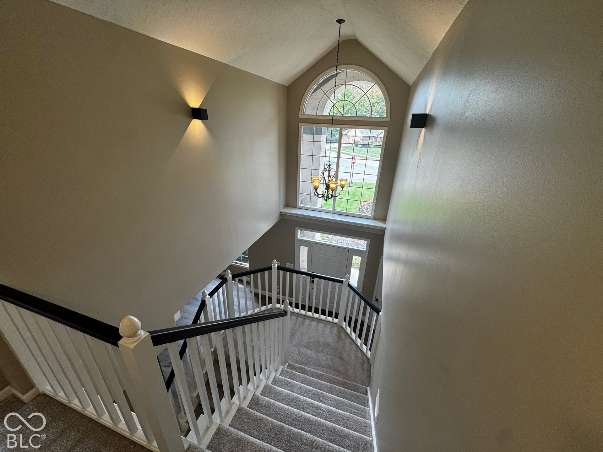 Two-story entryway with vaulted ceiling, arched window, chandelier, and a carpeted staircase with white balusters and dark handrails
