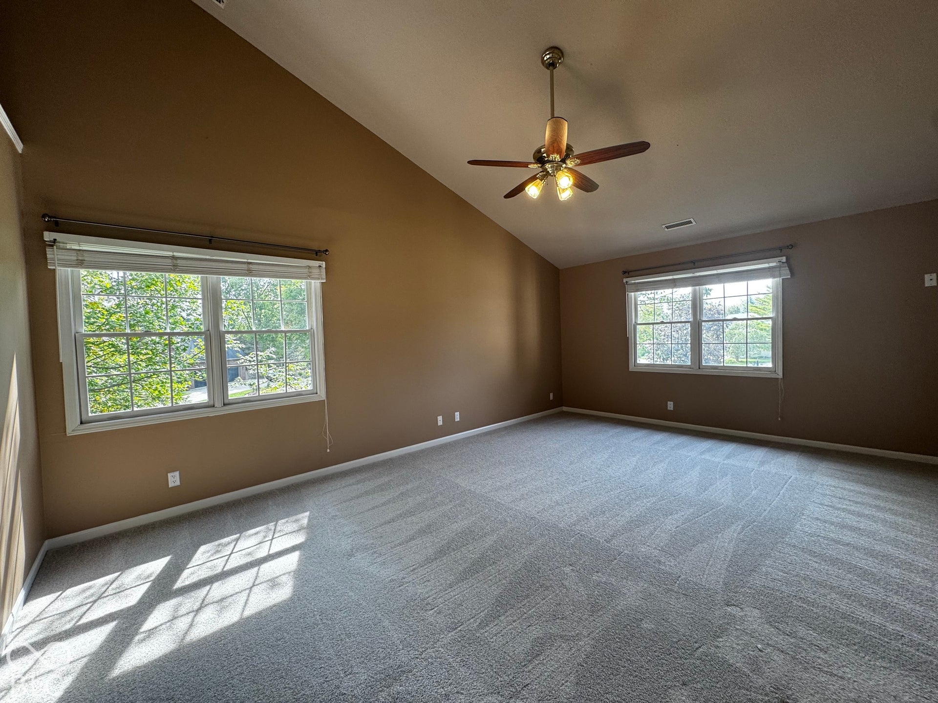 Vaulted ceiling, two large windows, neutral-toned walls, ceiling fan with integrated lighting, and gray carpeting