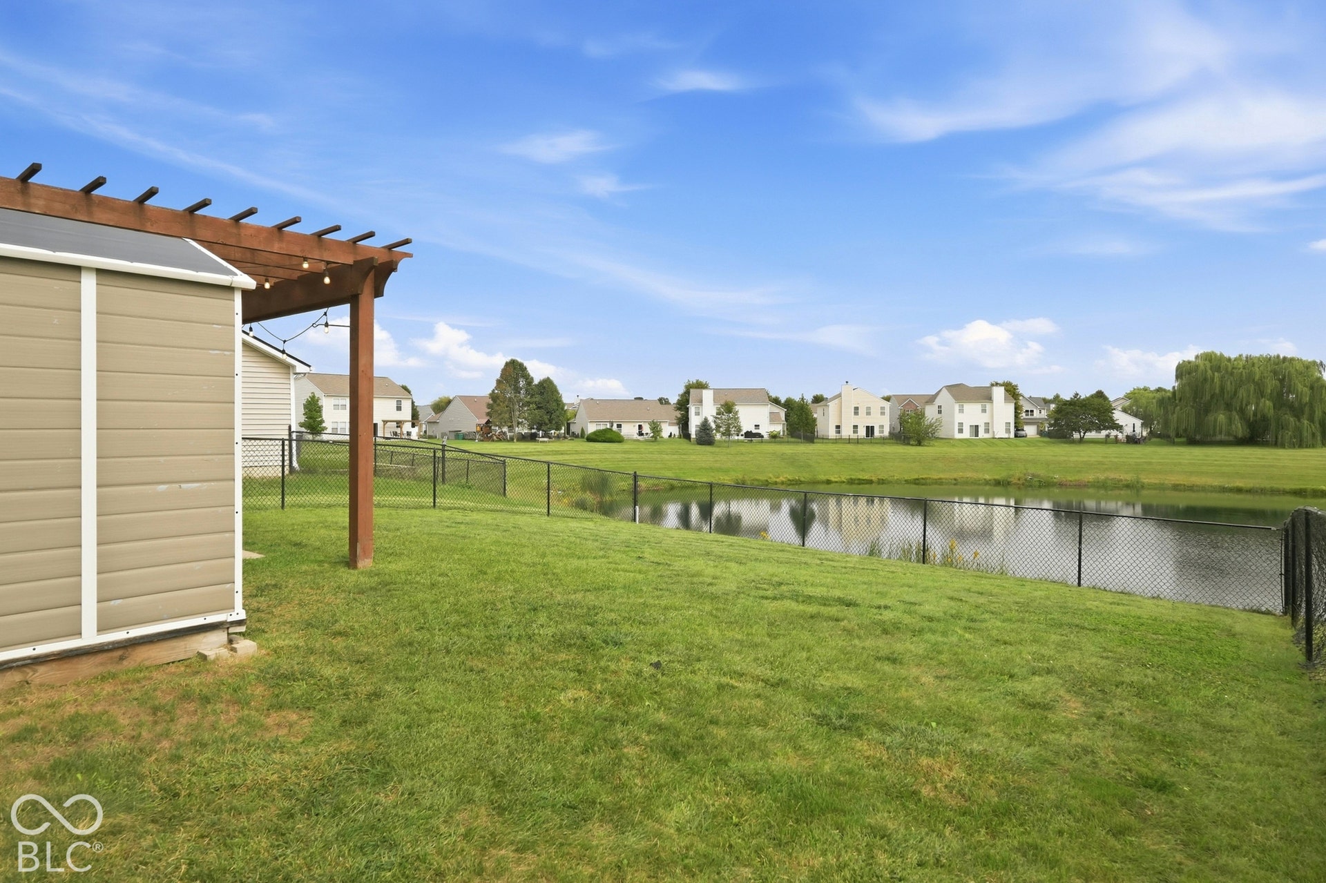 Manicured lawn with a pergola, a storage shed, and a chain-link fence bordering a pond