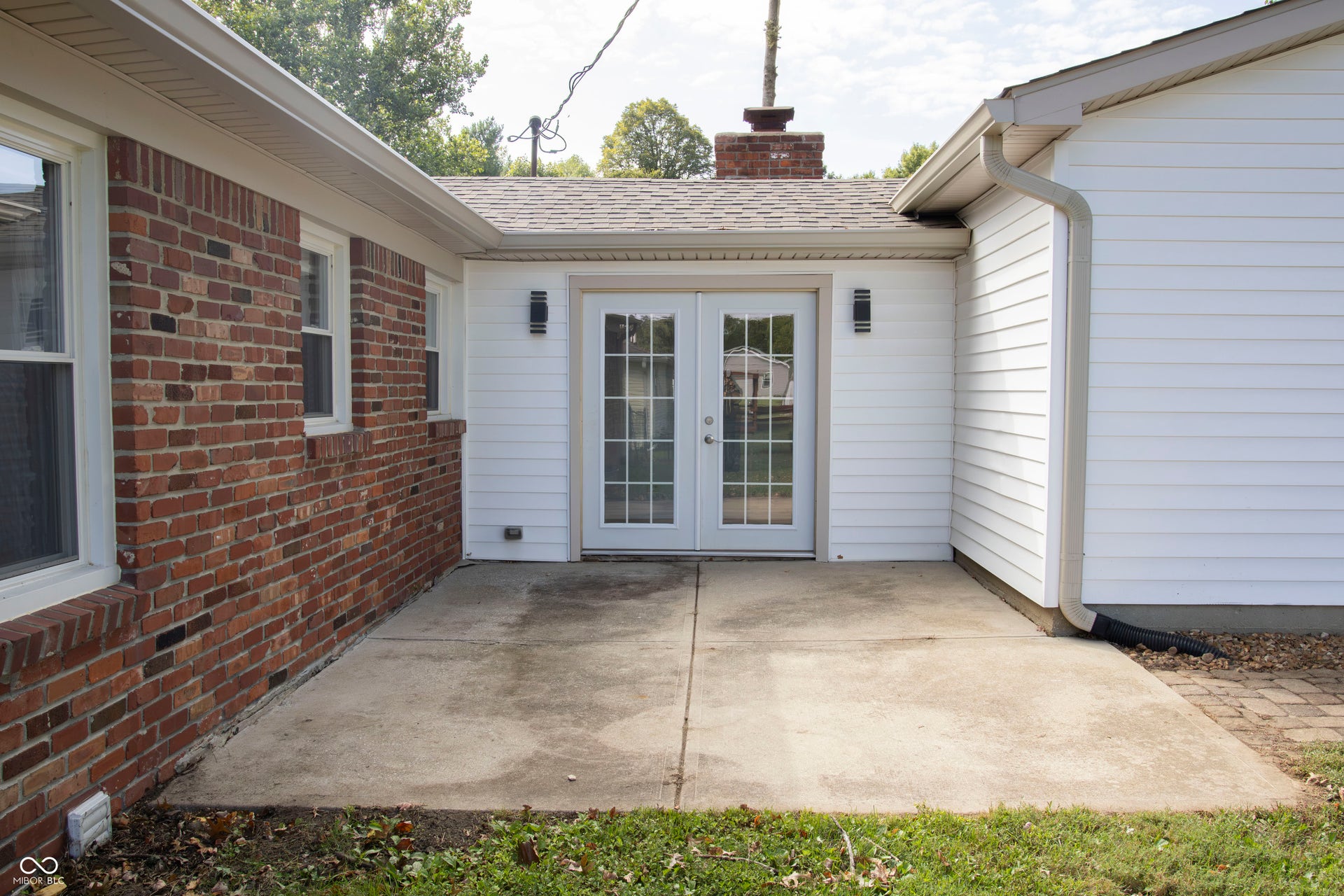 Exterior patio featuring concrete slab, brick siding, white vinyl siding, double French doors with grid panes, and exterior sconce lighting