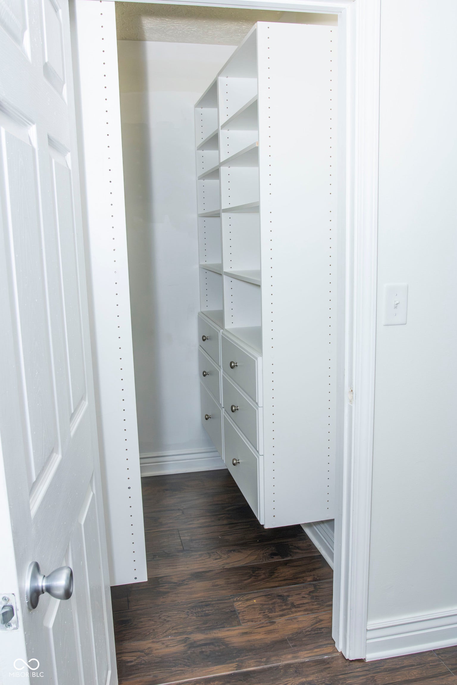 Integrated white shelving system with multiple adjustable shelves and built-in drawers, dark wood-finish flooring, and white trim