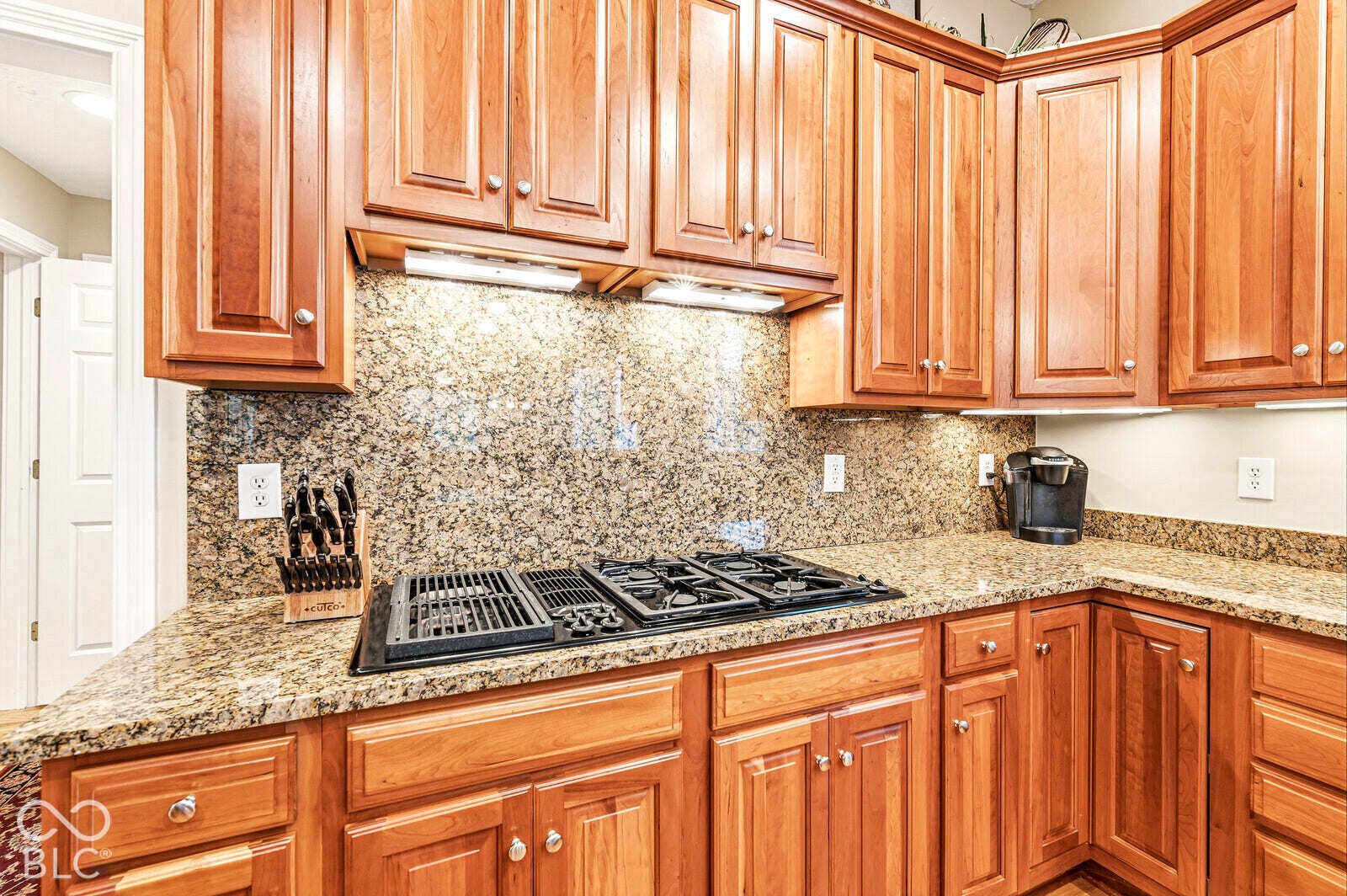 Wood cabinetry with recessed panel doors, under-cabinet lighting, a built-in gas range, and speckled stone countertops with a matching backsplash