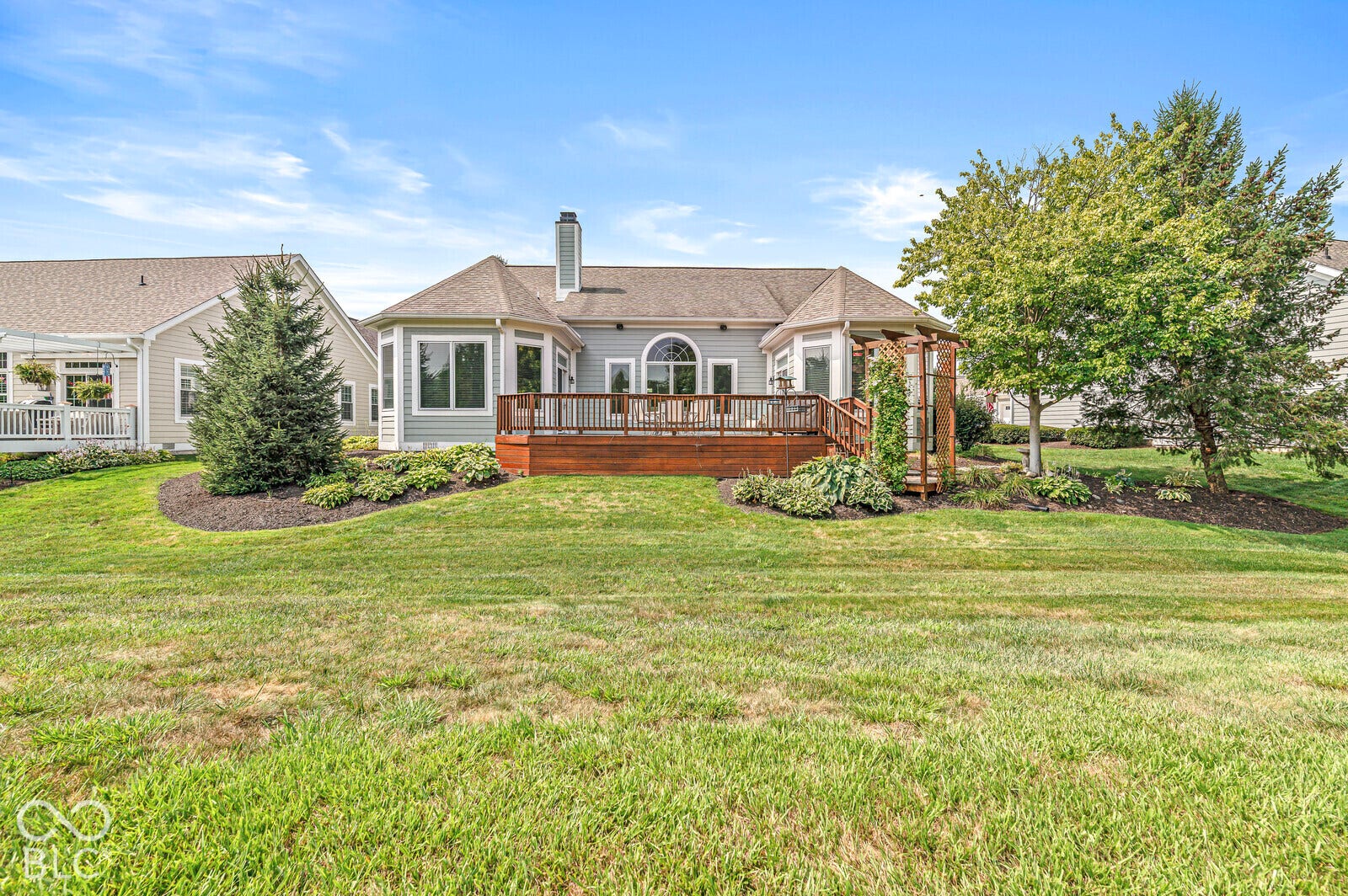 Rear exterior featuring a wood-finish deck, multiple large windows, and a landscaped perimeter