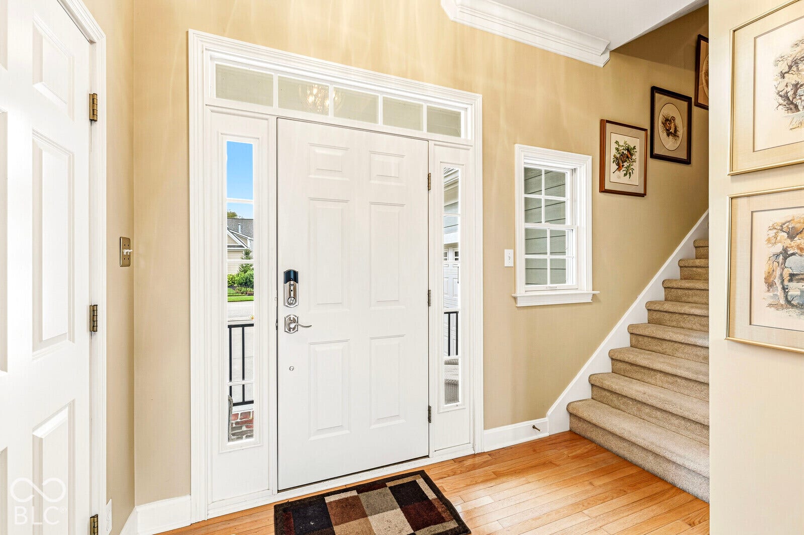 Hardwood flooring, paneled front door with sidelights and transom window, crown molding, and a carpeted staircase with white risers