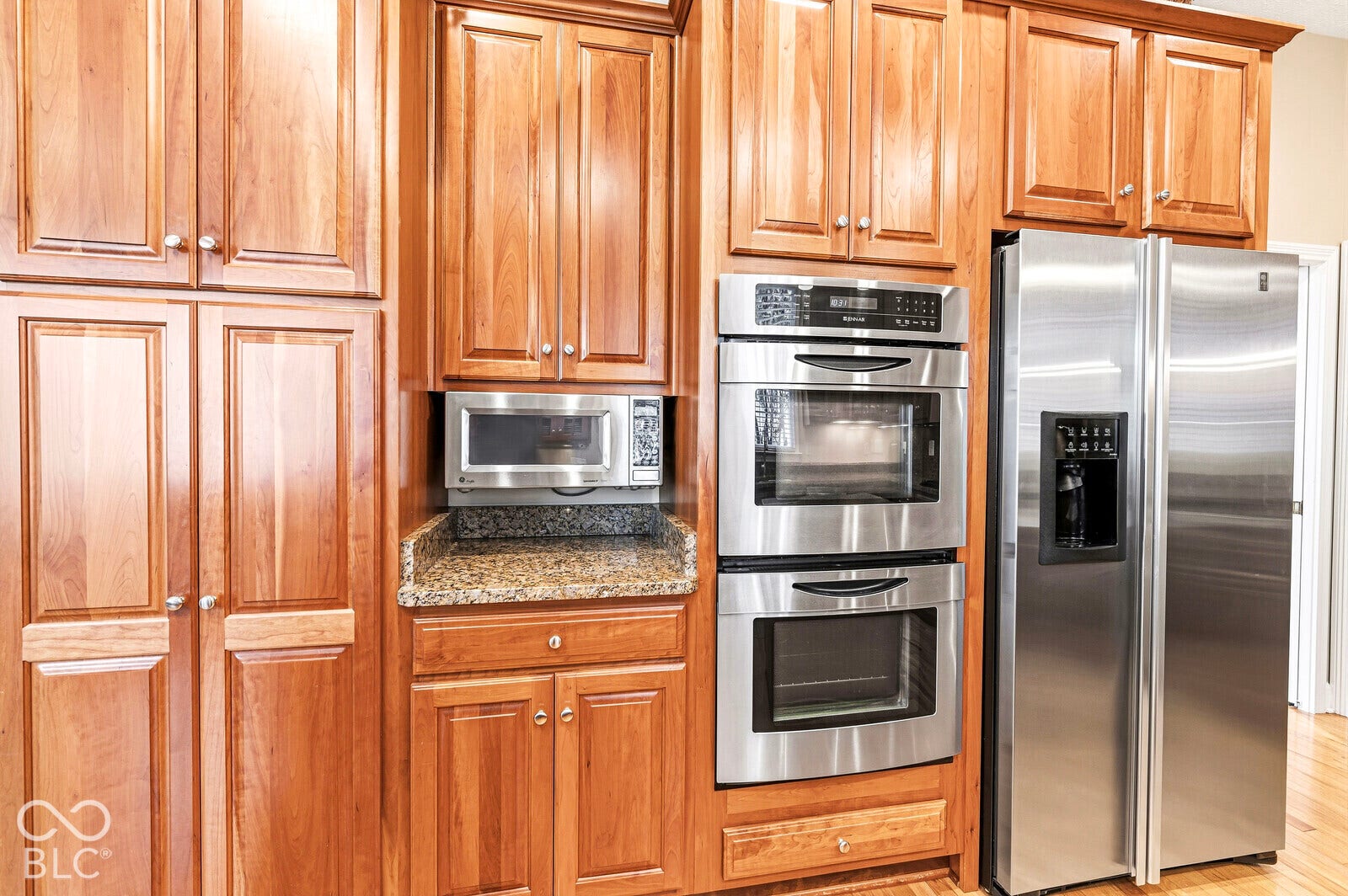 Built-in stainless steel double ovens, stainless steel microwave, stainless steel side-by-side refrigerator, granite-look countertop, and wood cabinetry with crown molding
