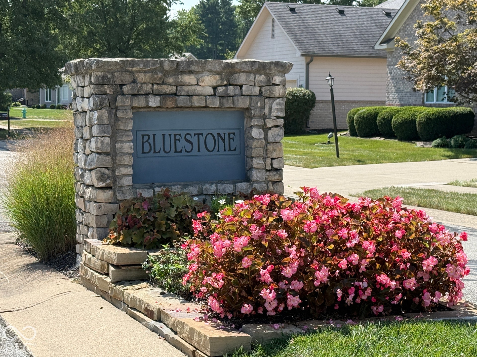 Stone-clad monument sign with "BLUESTONE" inscription, surrounded by manicured landscaping, vibrant flowering plants, and ornamental grasses welcoming you and your guests.