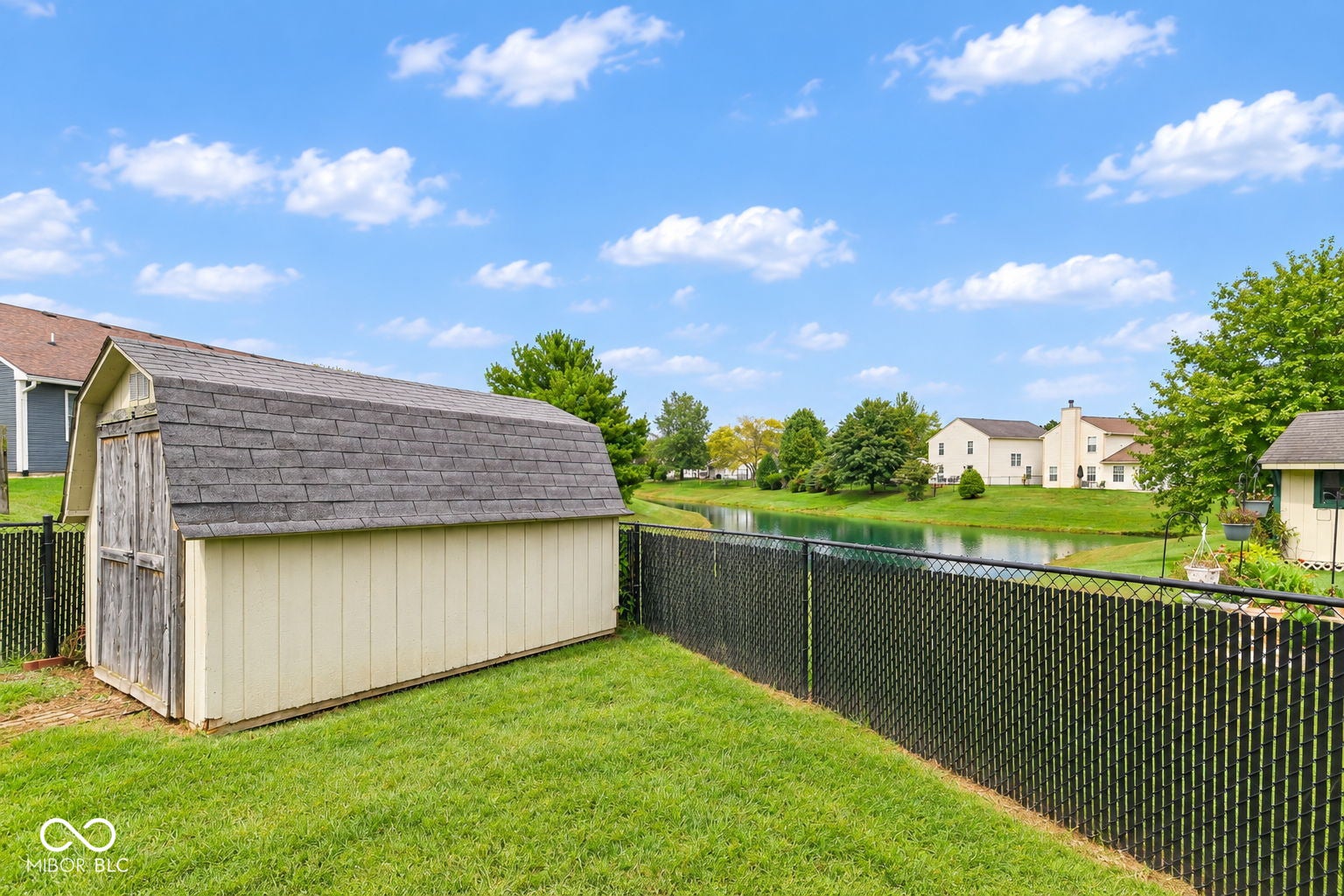 Expansive lawn with a black chain-link fence, a storage shed with an asphalt shingle roof, and a pond