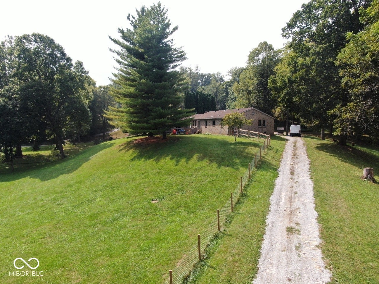 Gravel driveway leading to a brick exterior residence with a fenced perimeter and mature trees
