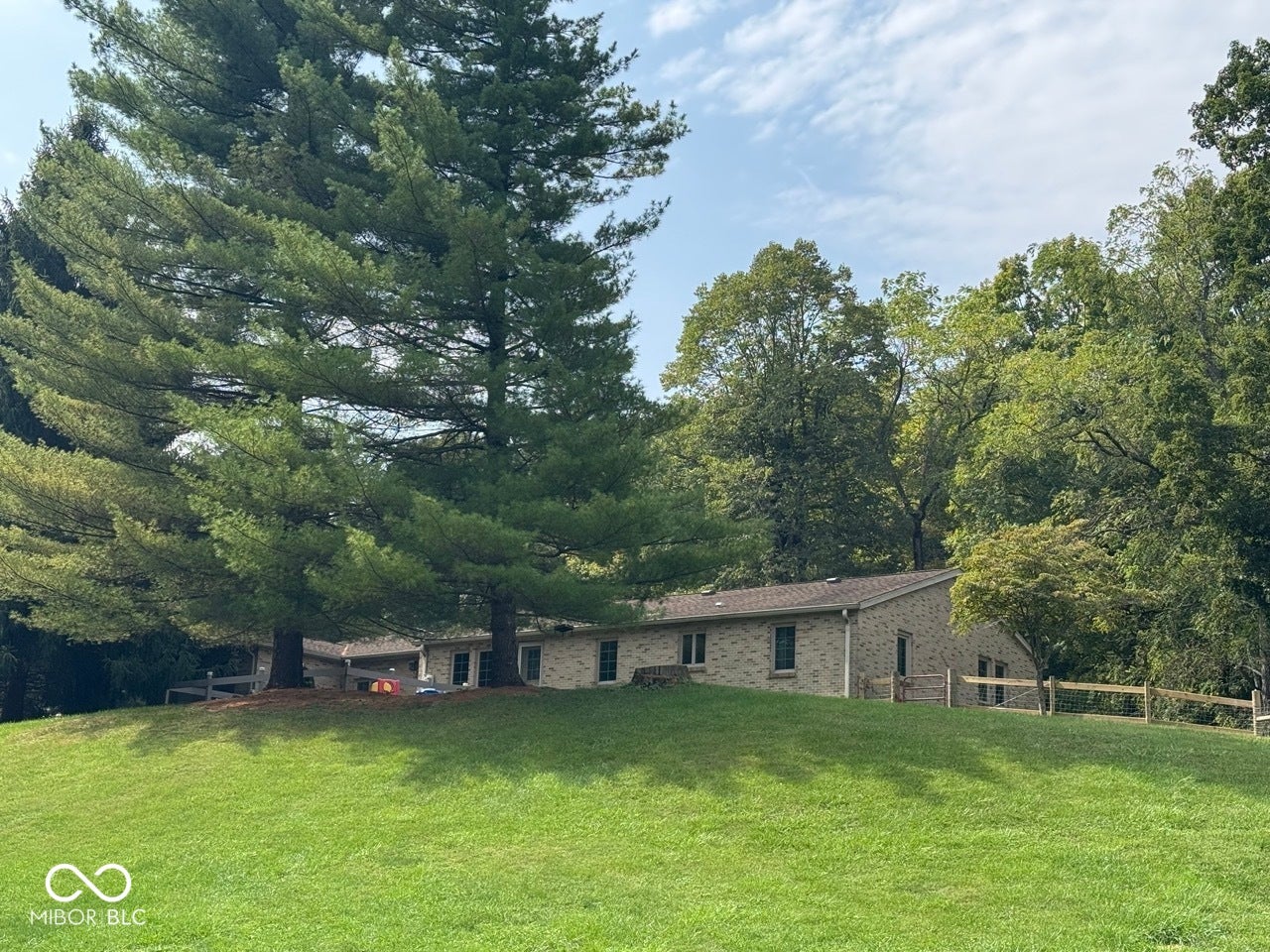 Expansive grassy hillside leading to a stone-clad residence with a shingled roof, bordered by a rustic wooden fence and mature trees