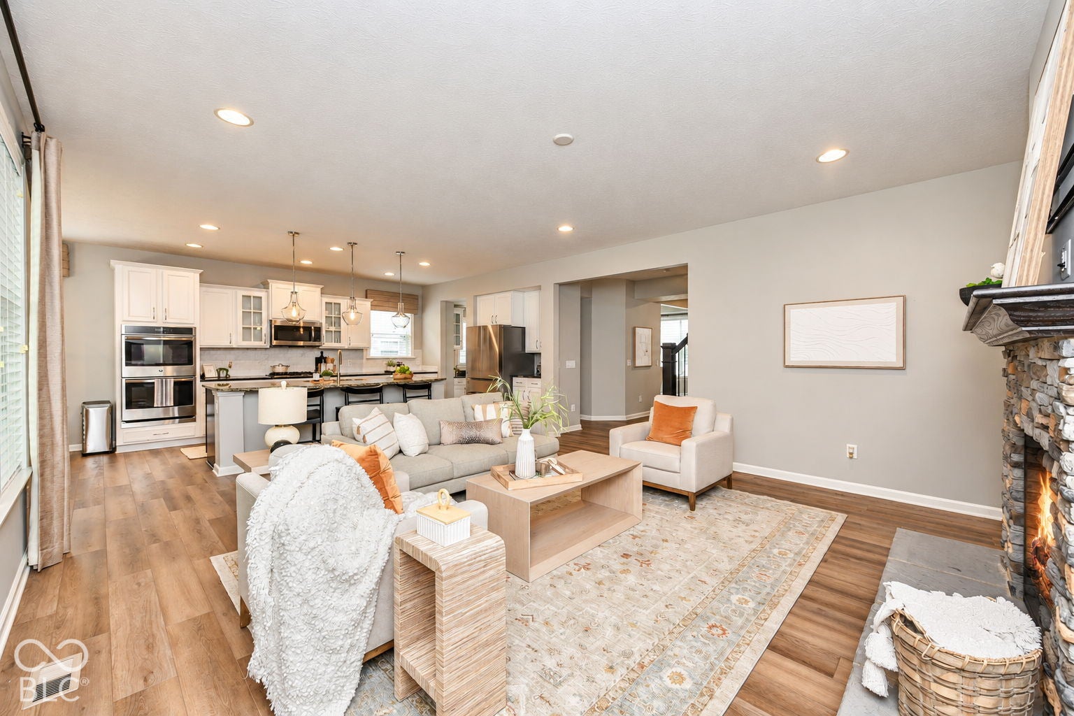 Open-concept living space featuring wood-finish flooring, a stone fireplace with a hearth, recessed lighting, and a kitchen with white cabinetry and stainless steel double ovens