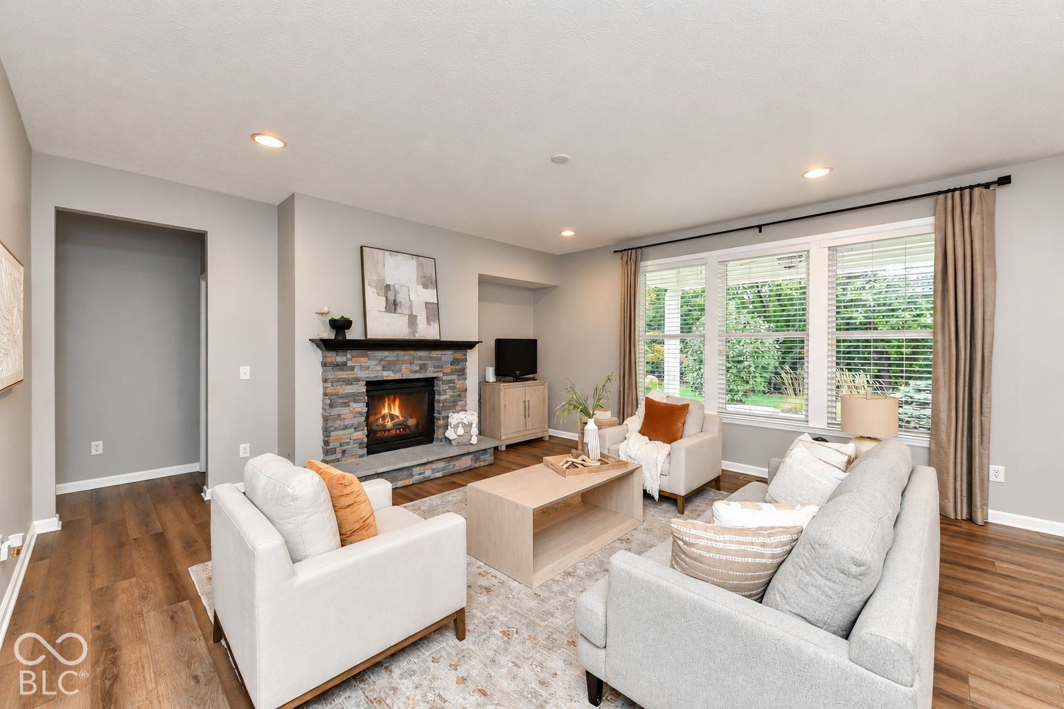 Stacked stone fireplace with a dark wood mantle, recessed lighting, wood-finish flooring, and multiple windows