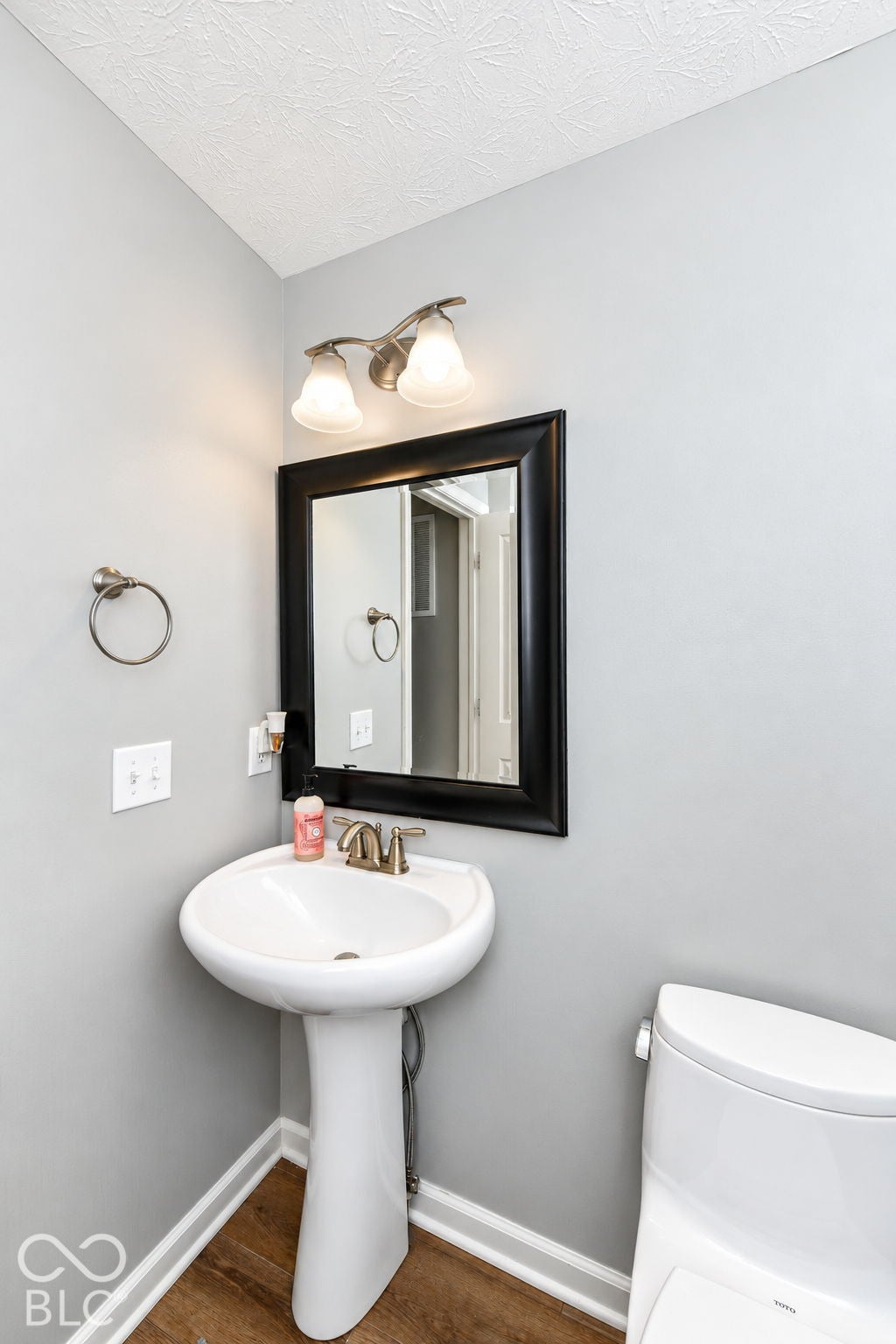 Pedestal sink with brass-finish faucet, framed mirror with two-bulb vanity light, and wood-finish flooring