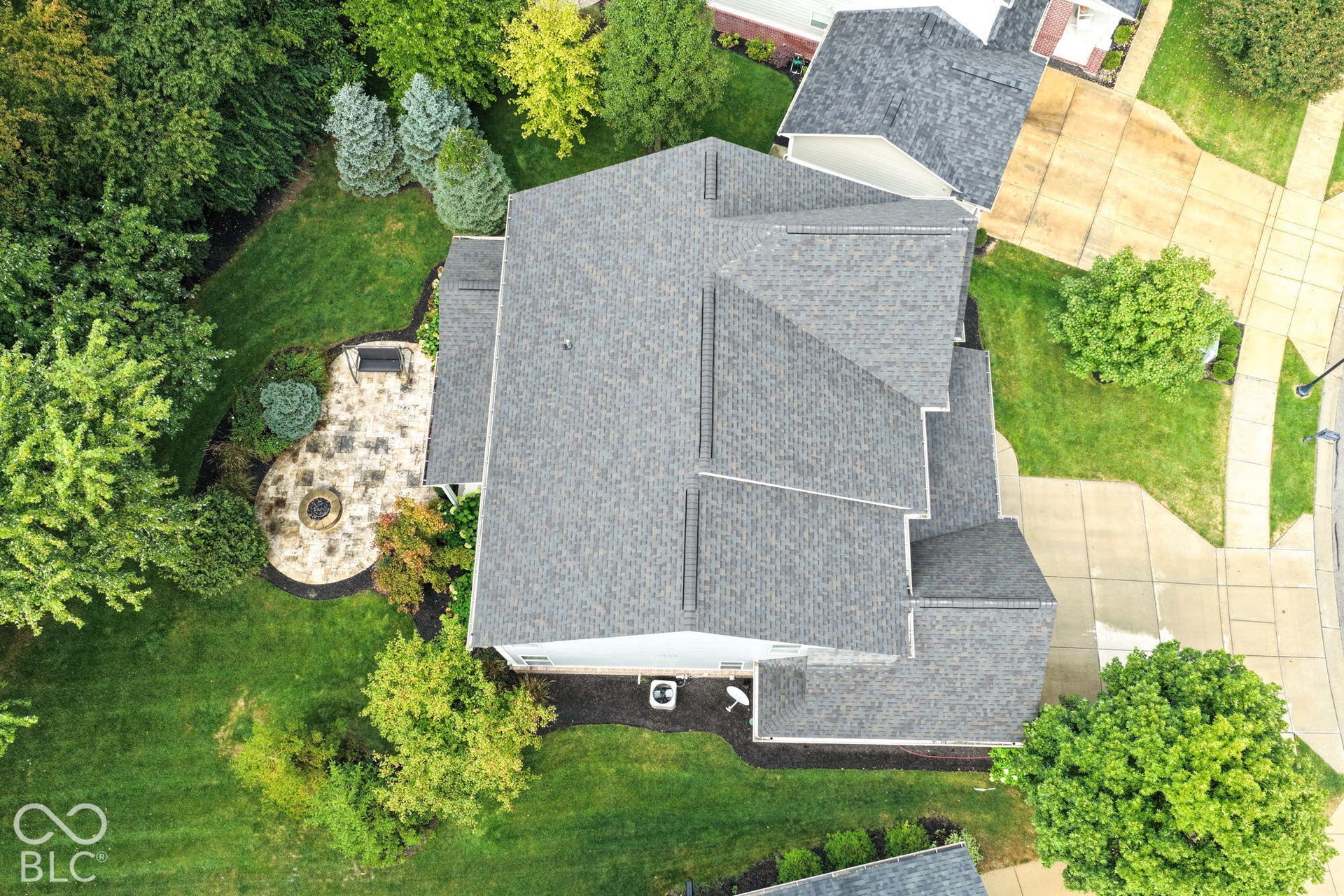 Pitched shingle roof, well-maintained lawn, stone patio with built-in fire pit, mature perimeter landscaping, concrete driveway