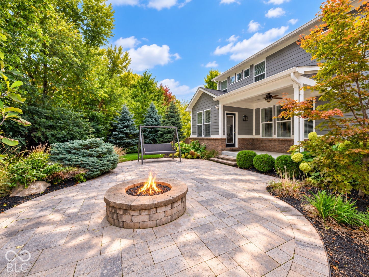 Paver patio with a stone fire pit, mature landscaping, and a covered porch with a ceiling fan