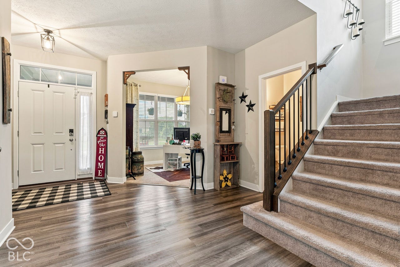 Wood-finish flooring, two-panel entry door with transom window, carpeted staircase with dark wood newel post and metal balusters, and recessed archway
