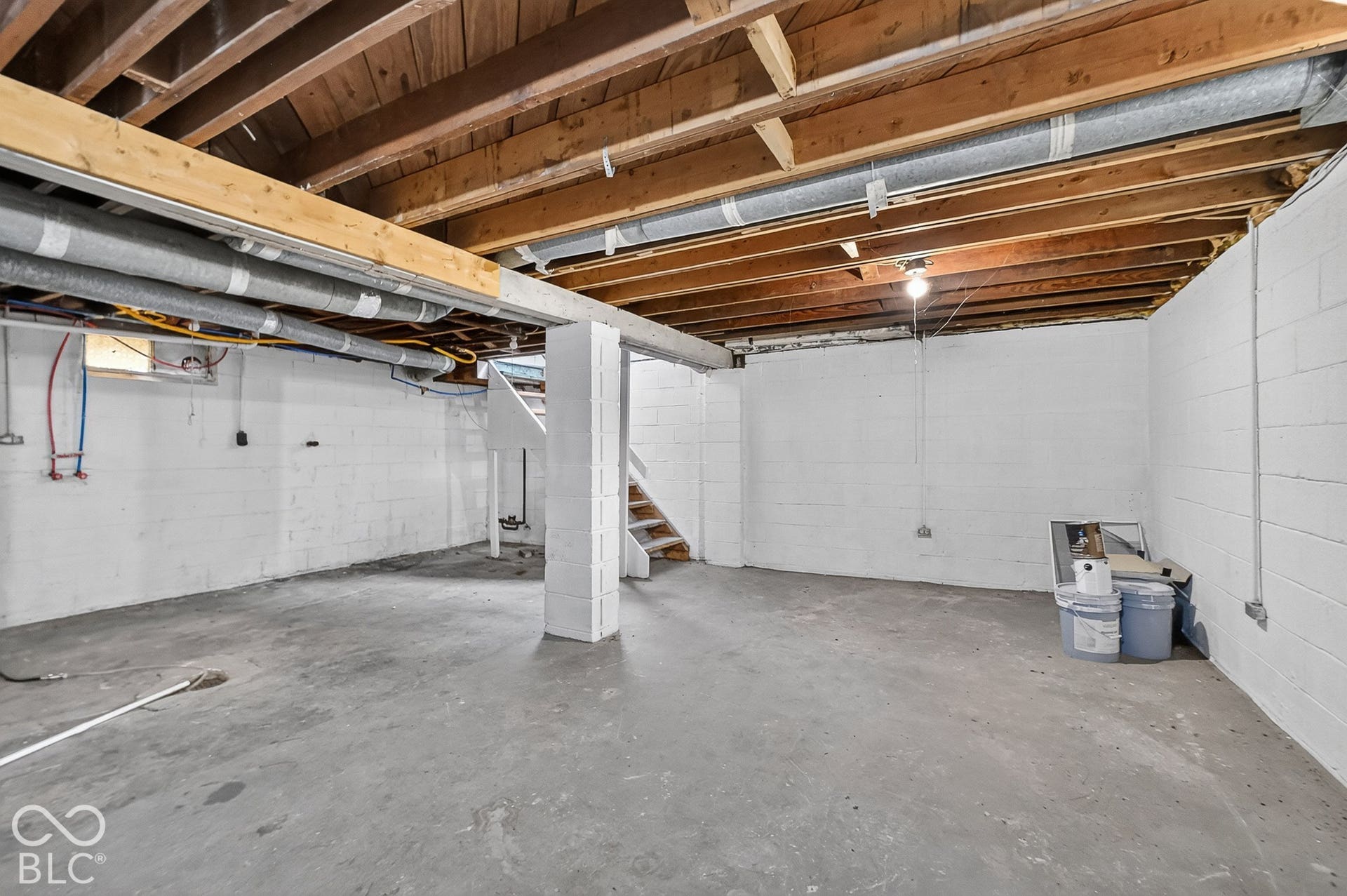 Exposed wood ceiling joists with visible ductwork, concrete flooring, painted cinder block walls, and open wooden stairs