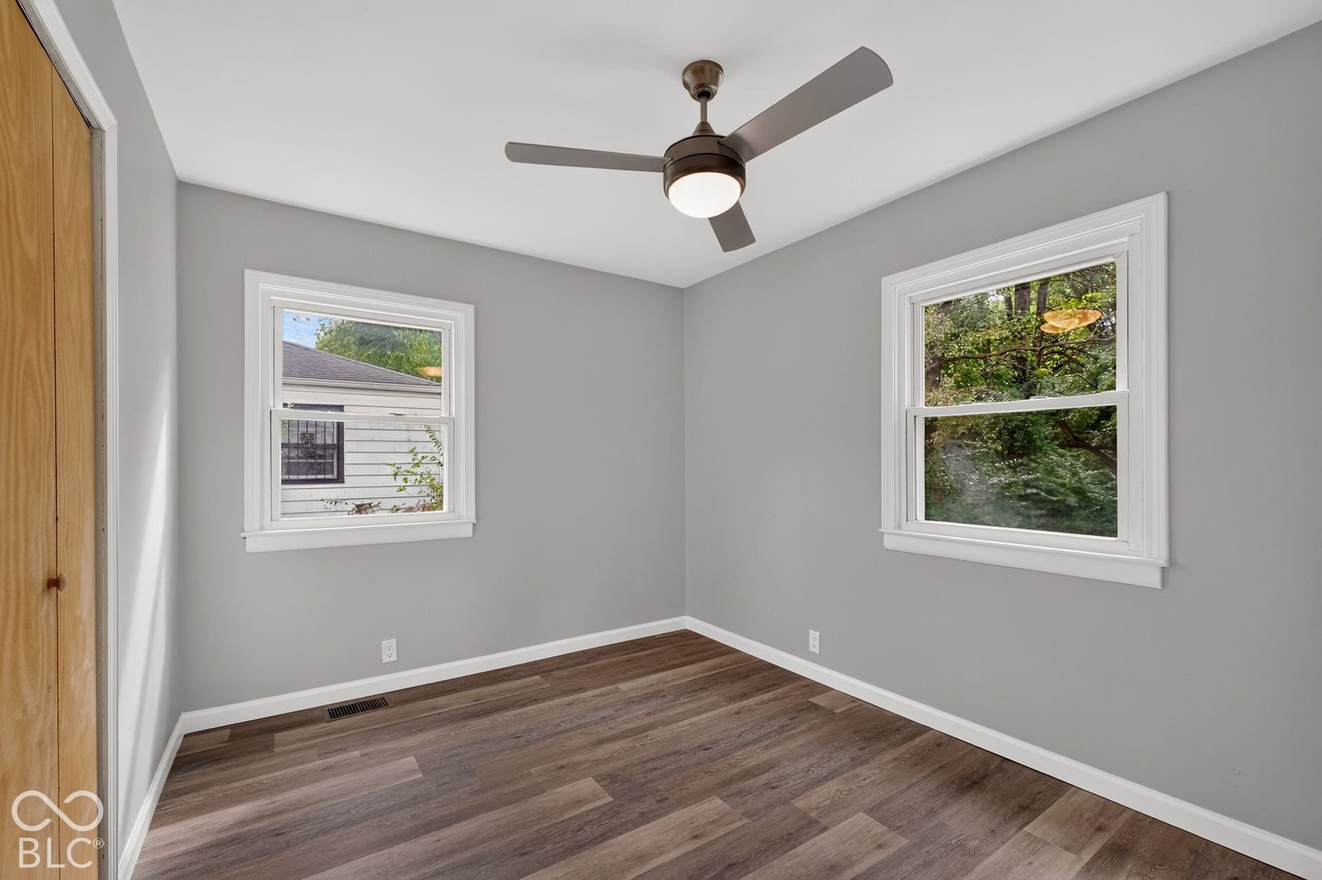 Wood-finish flooring, two windows with white trim, a ceiling fan with integrated lighting, and light gray wall paint