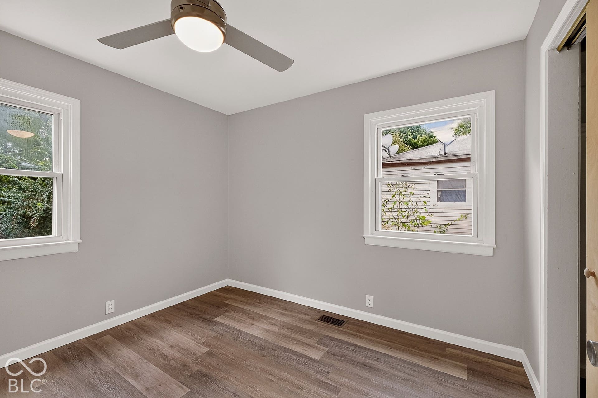 Wood-finish flooring, two white-trimmed windows, a ceiling fan with integrated lighting, and light gray wall paint