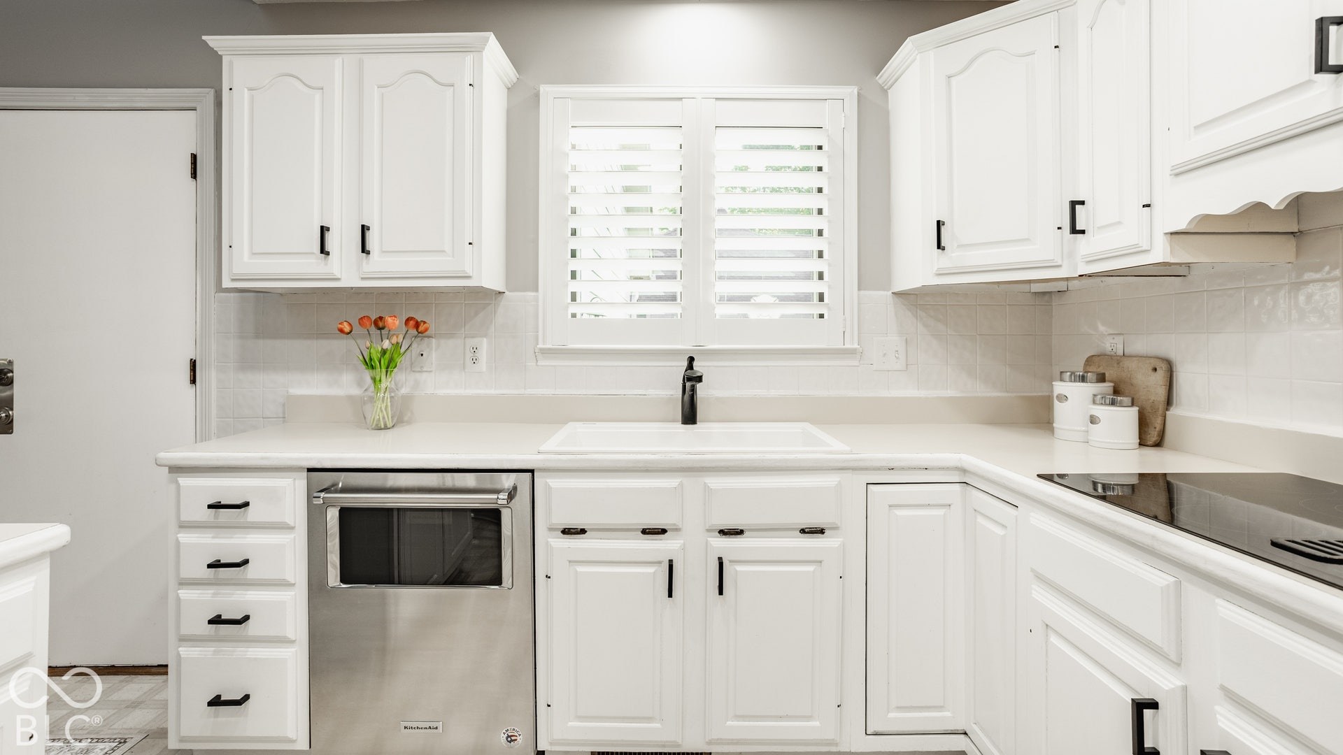 White cabinetry with black hardware, integrated electric cooktop, stainless steel dishwasher, white countertop, and white tile backsplash
