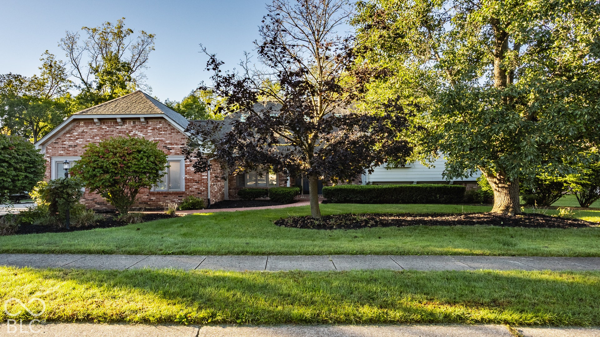 Brick home with gabled roofline, mature trees, manicured lawn, and paved sidewalk