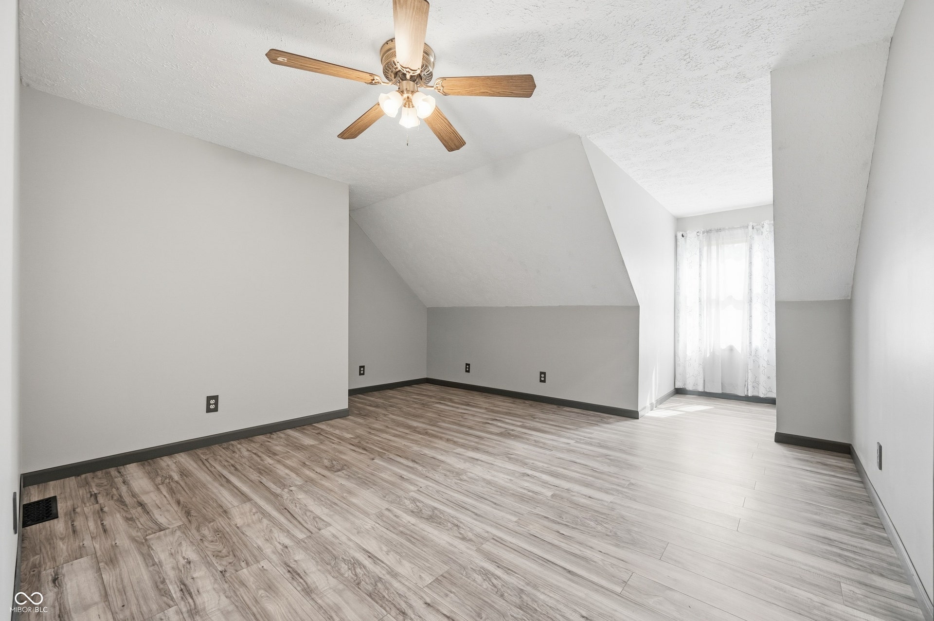 Sloped ceilings with textured finish, wood-finish flooring, ceiling fan with integrated lighting, and a prominent window