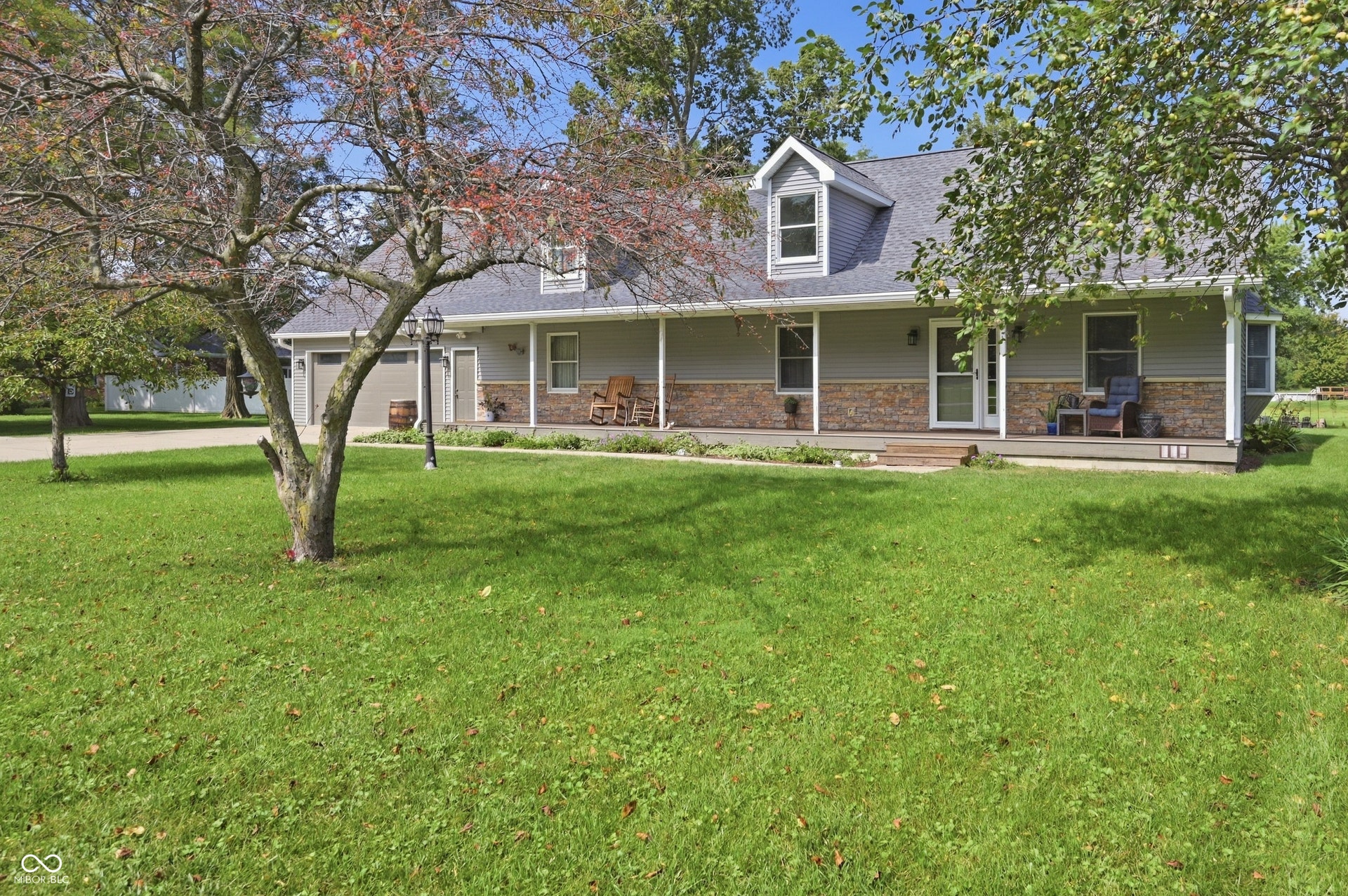 Expansive front lawn with mature trees, covered front porch, dormer windows, attached garage, and stone facade accents