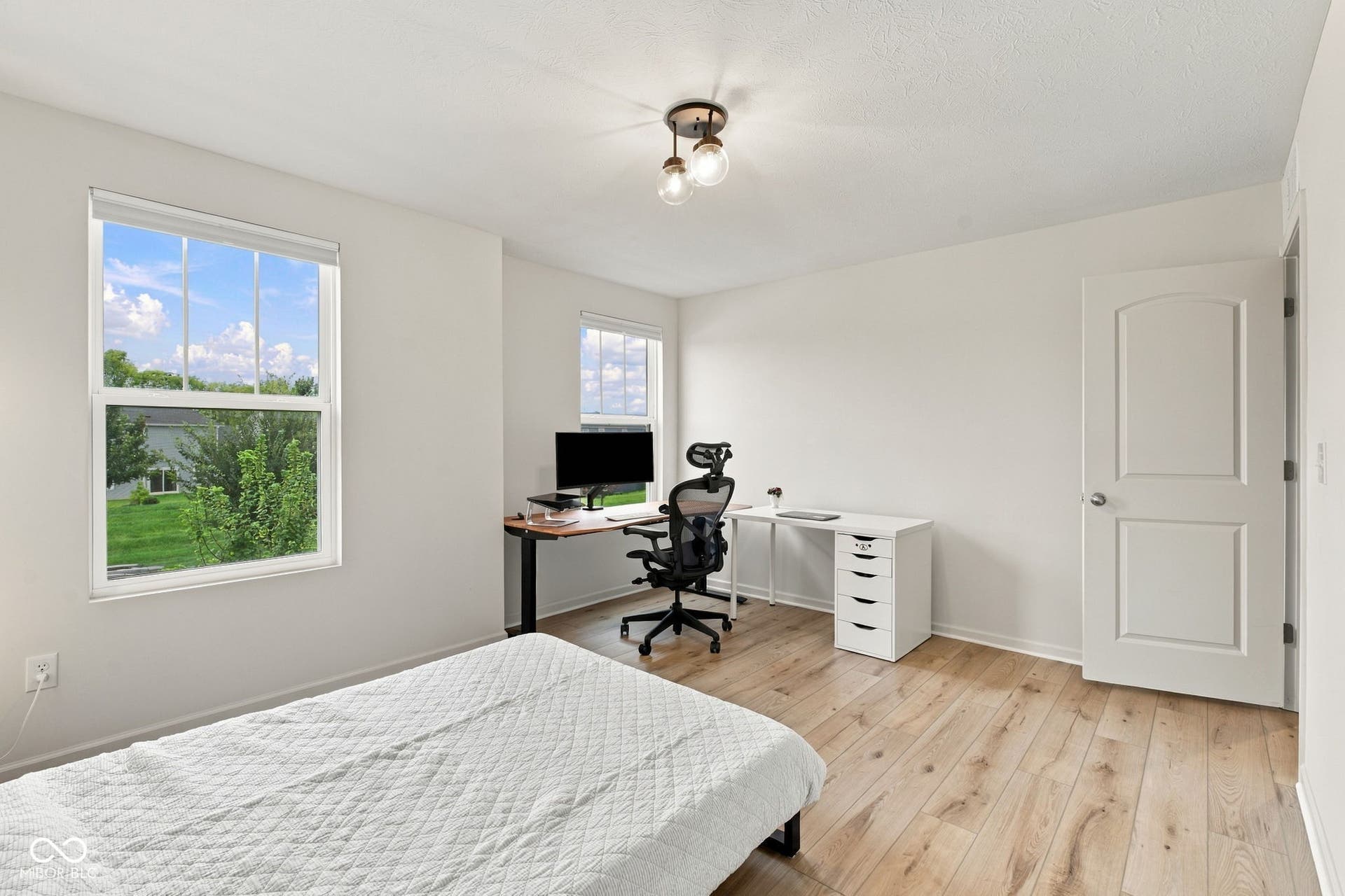 Light wood-finish flooring, two windows with white trim, a semi-flush mount ceiling light fixture, and a white interior door with an arched panel