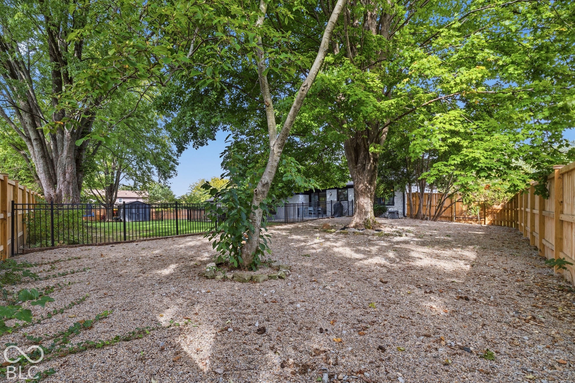 Expansive gravel yard with mature trees, bordered by a wood privacy fence and a black metal fence