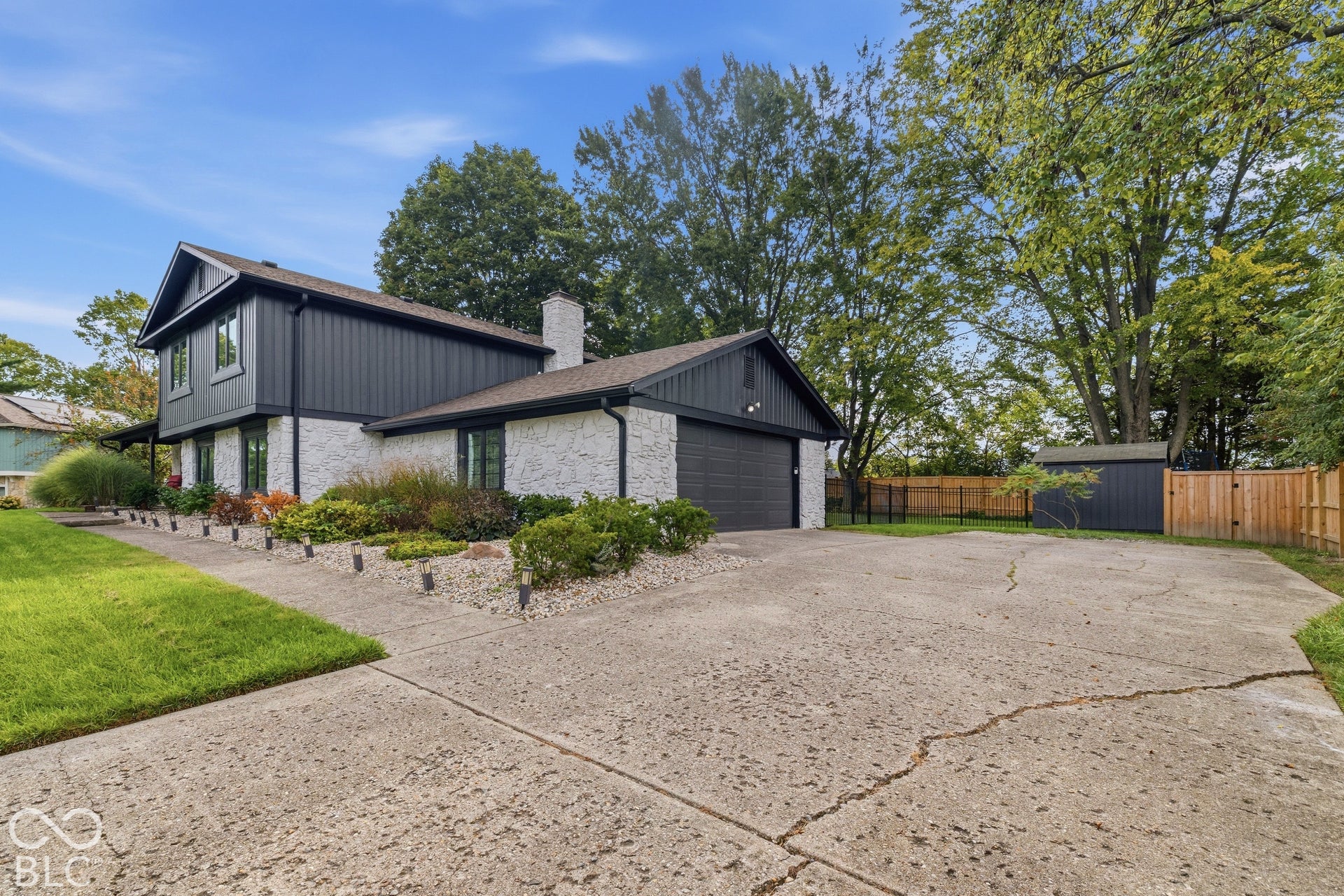 Contemporary exterior with stone and vertical siding, attached garage, dedicated parking area, mature trees, and a storage shed