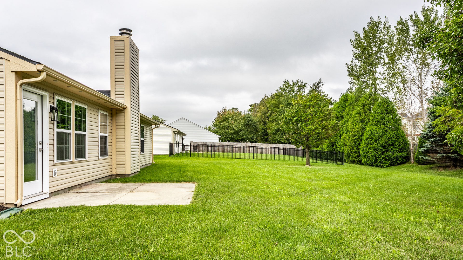 Expansive backyard with a concrete patio, mature trees, and a black metal fence