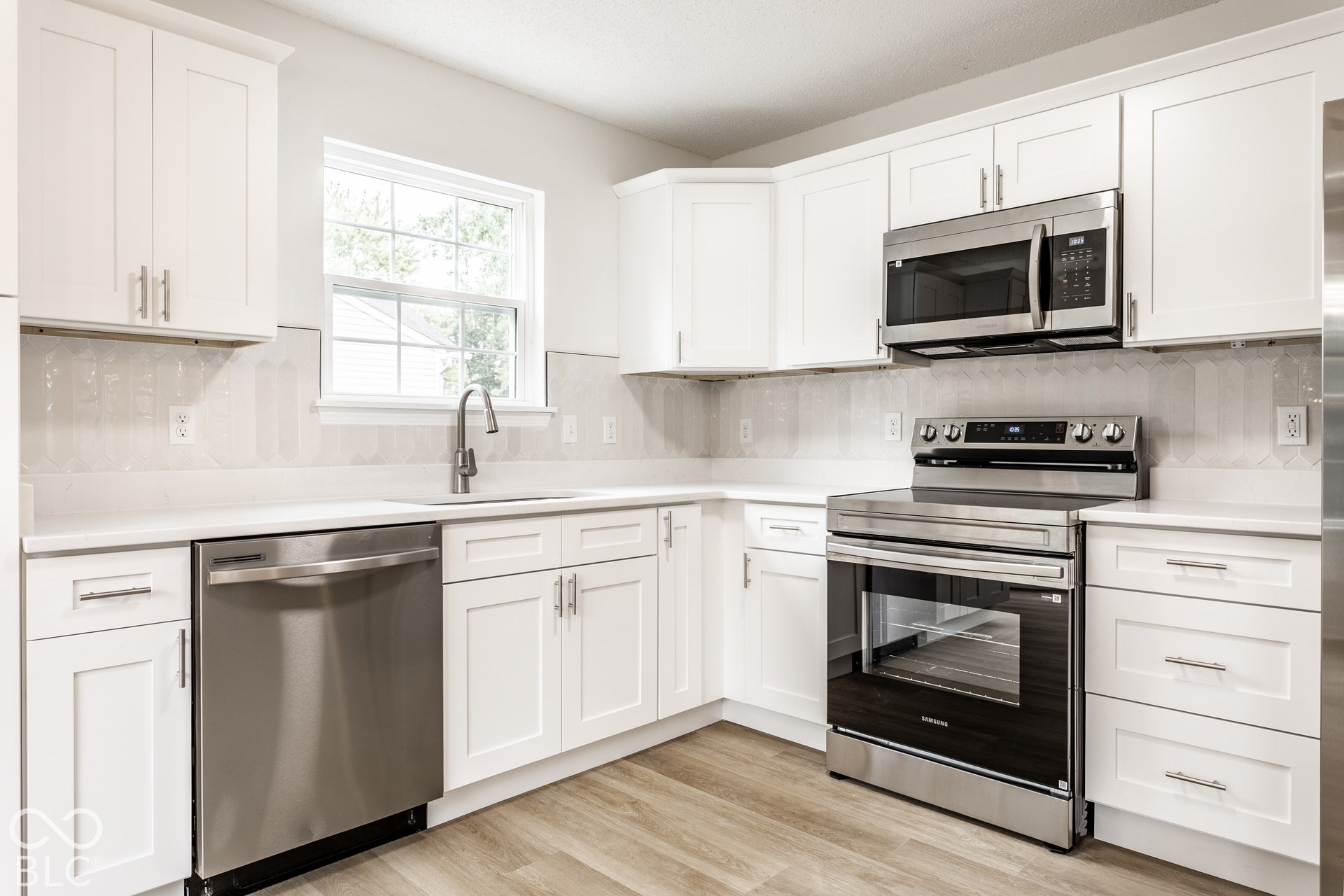 White shaker-style cabinetry with stainless steel hardware, light-toned countertops, tile backsplash, stainless steel appliances, and wood-finish flooring