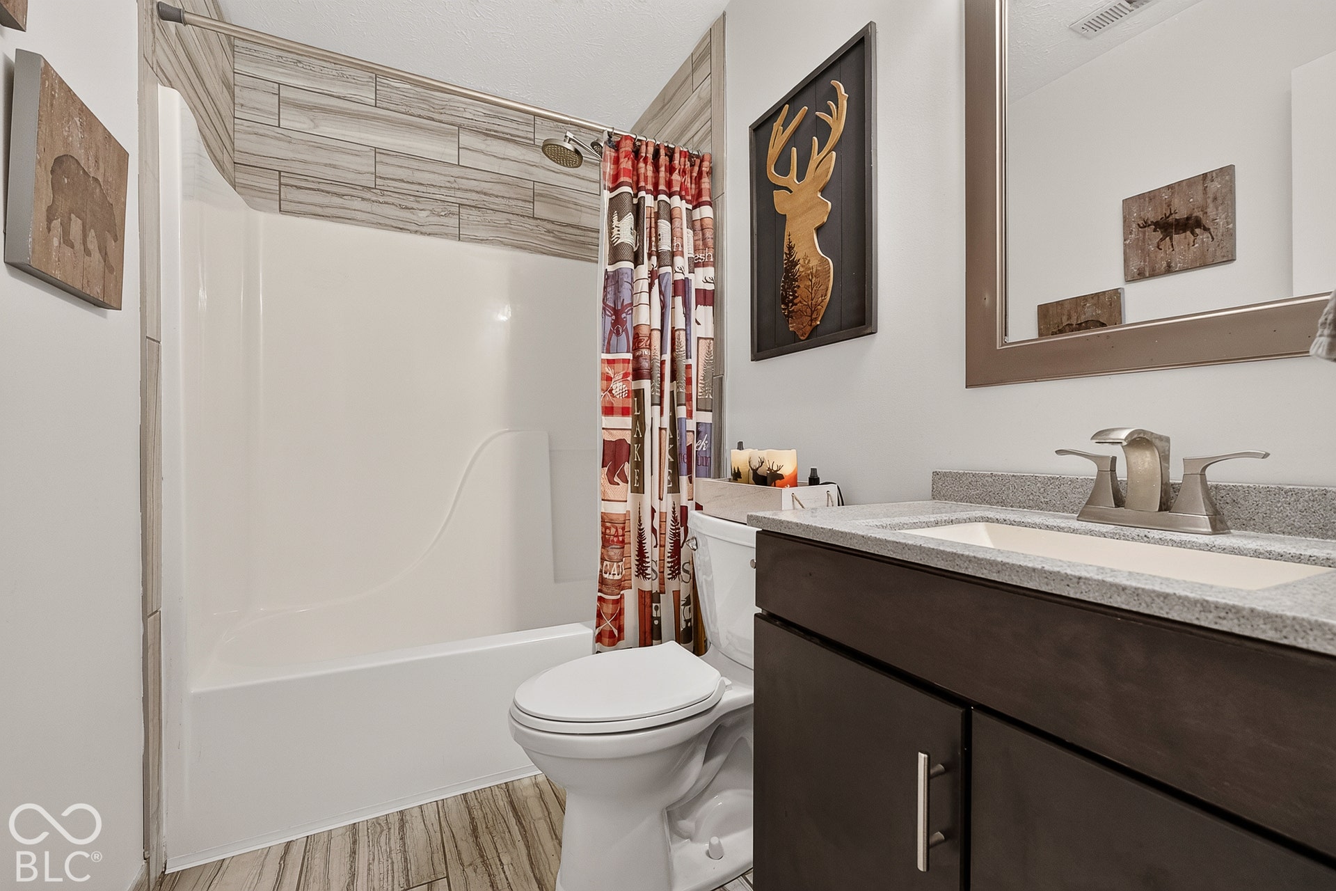 Integrated shower and tub unit with tiled surround, wood-finish flooring, dark wood vanity with a light-colored countertop, and a vanity mirror with a metallic frame