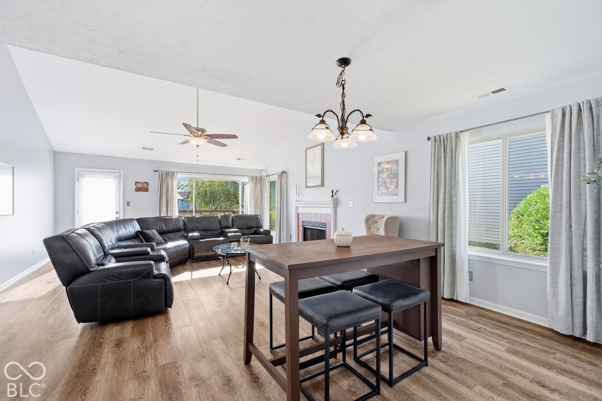 Vaulted ceiling with ceiling fan, wood-finish flooring, fireplace with white mantel, sliding glass door, and large window with window treatments