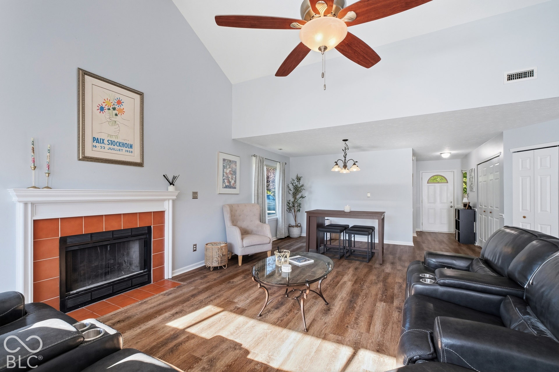 Vaulted ceiling with ceiling fan, wood-finish flooring, white mantel fireplace with terracotta tile surround, and a white front door with an arched glass insert