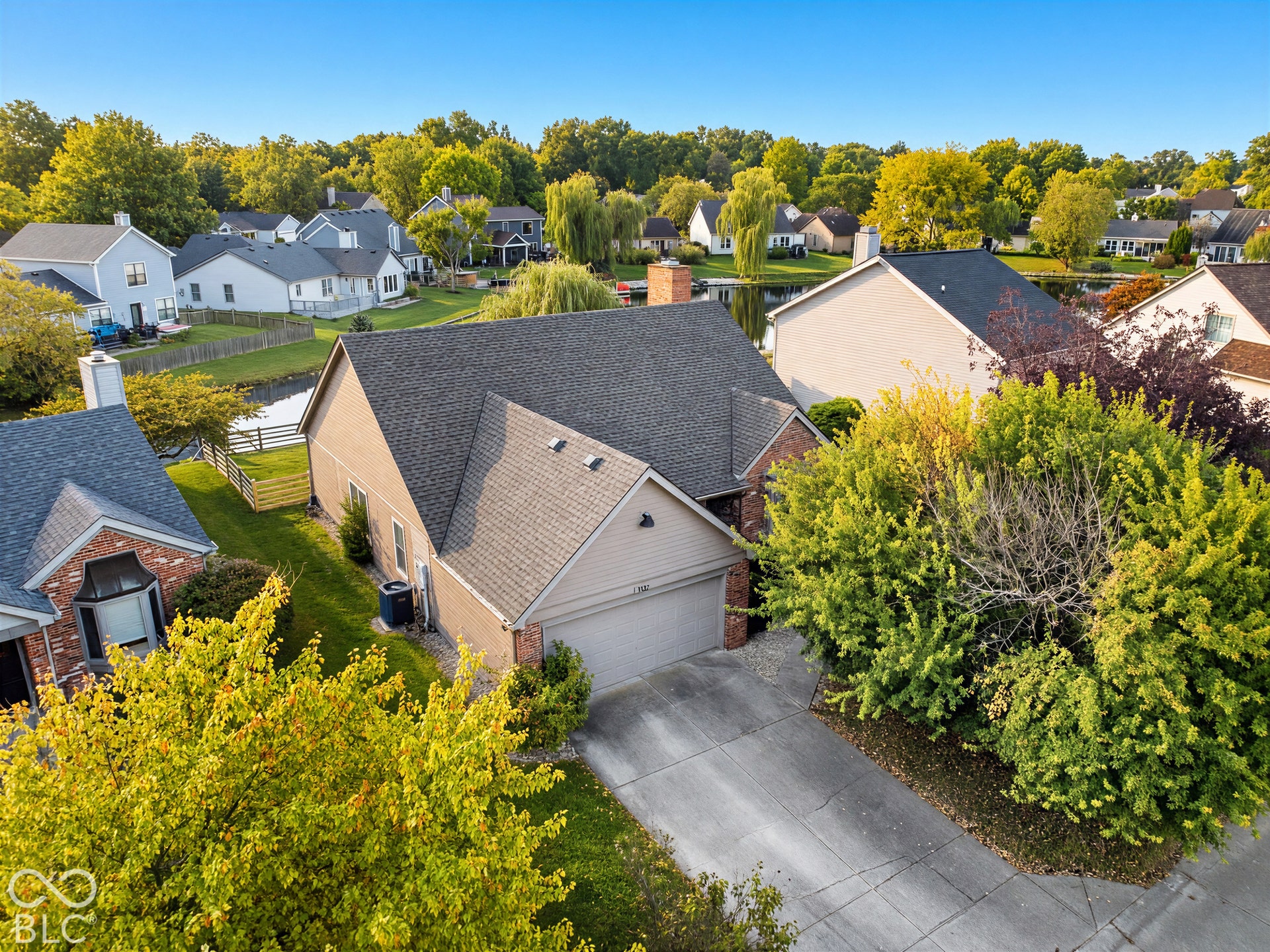 Asphalt shingle roofing, attached garage, concrete driveway, lush perimeter landscaping, and mature trees