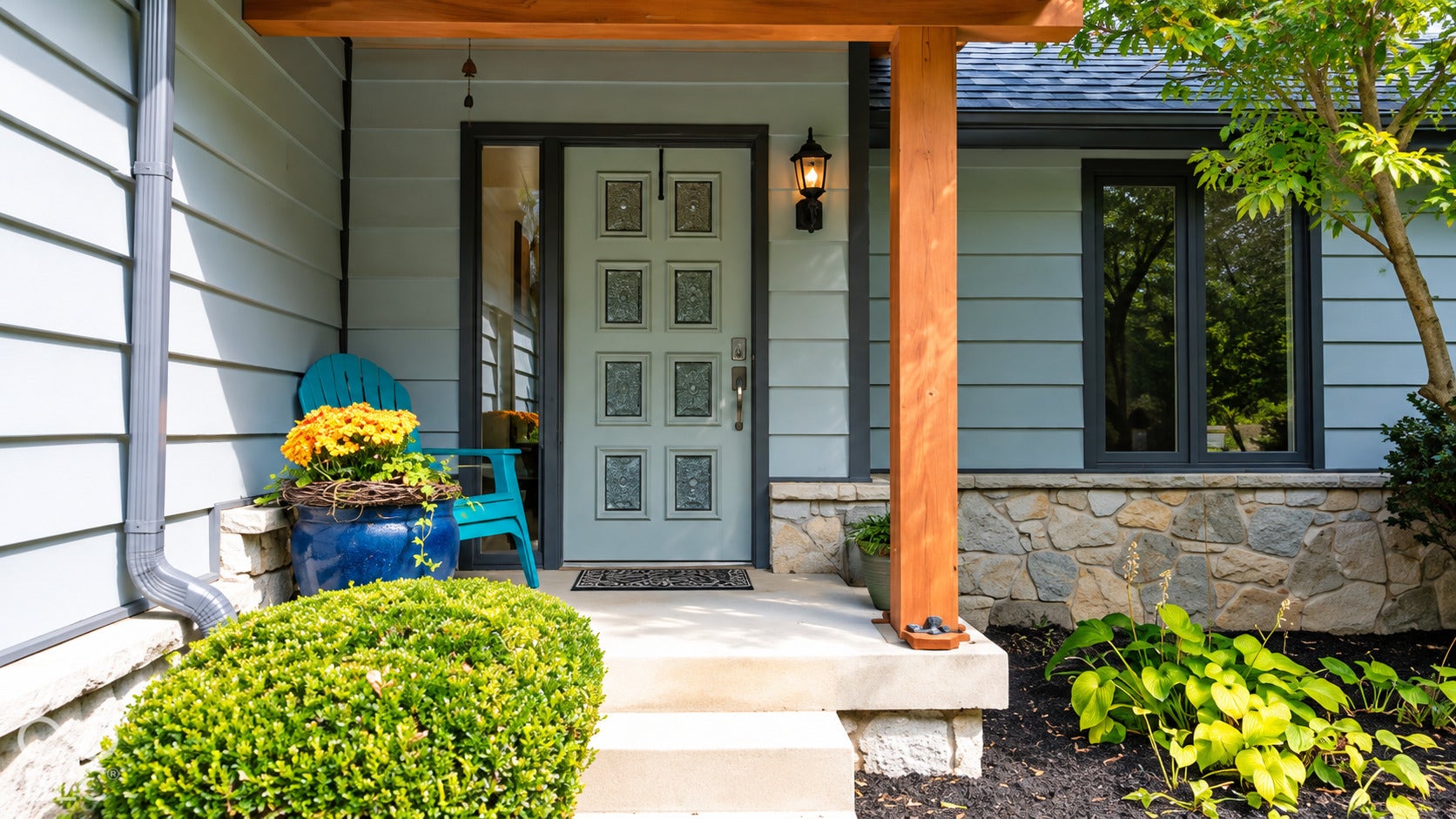Gable roofline with dark shingles, light blue horizontal cedar siding, natural stone foundation, wood support beams, and a panel-style front door with decorative glass inserts