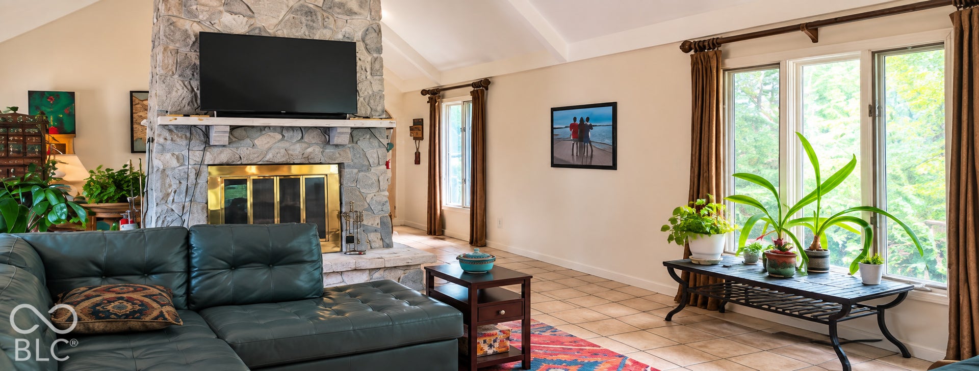 Vaulted ceiling, stone-clad fireplace with brass insert, large window with expansive glass, light-toned tile flooring, and neutral wall tones
