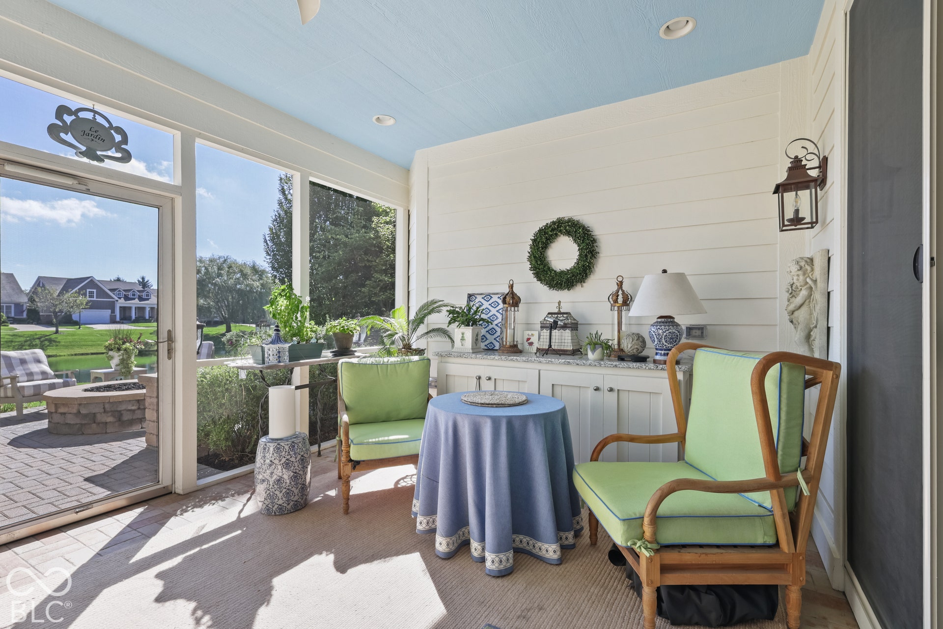 Virtually Altered - Screened porch featuring a light blue ceiling, white shiplap walls, a built-in white cabinet with a light-colored countertop, and a glass-paneled door opening to a paver patio with a stone fire pit