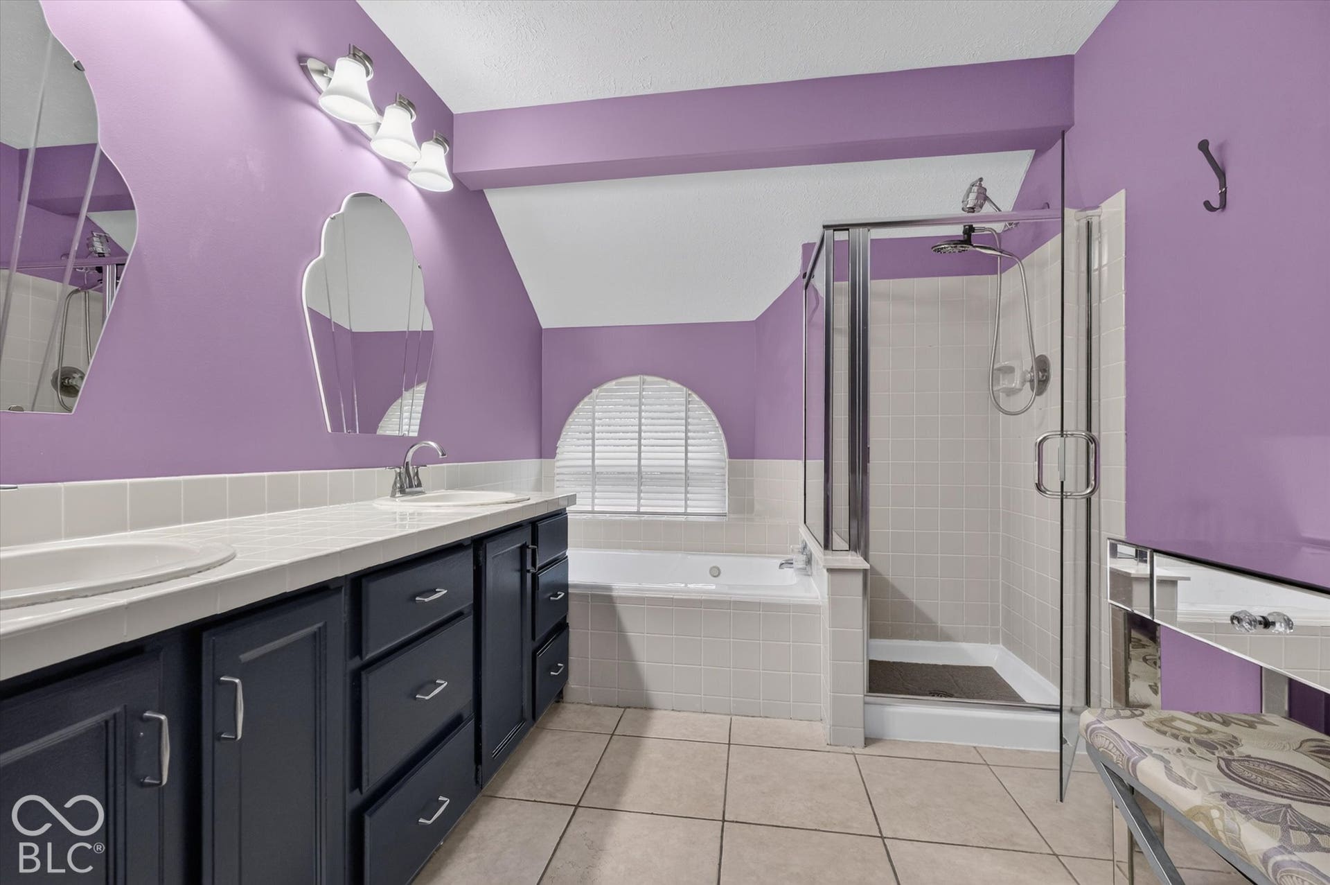 Dual vanity with undermount sinks and dark cabinetry, glass-enclosed shower, built-in soaking tub with arched window, and tile flooring