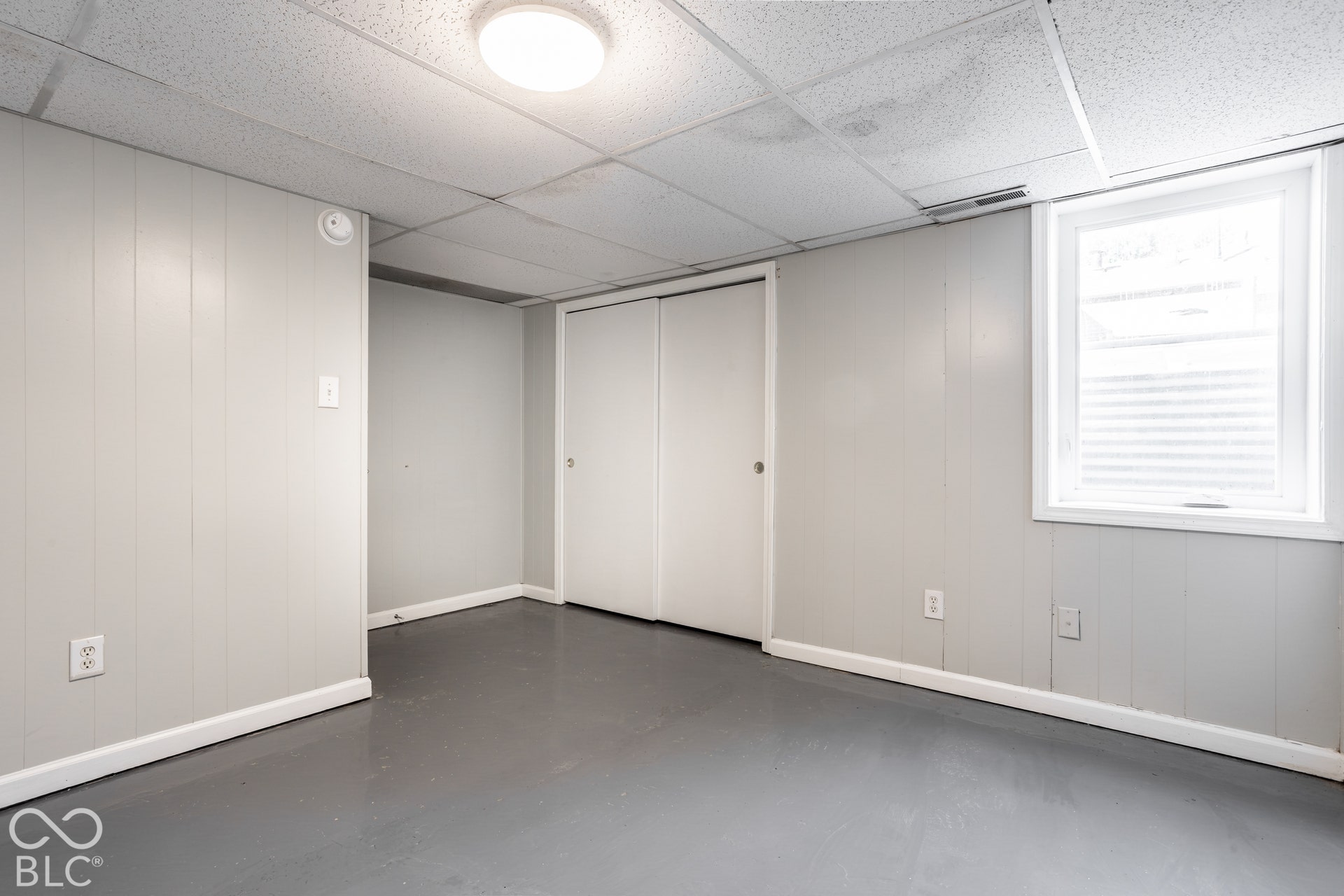Basement bedroomLight gray wall paneling, drop ceiling, flush-mount ceiling light, egress window with blinds, and bi-fold closet doors