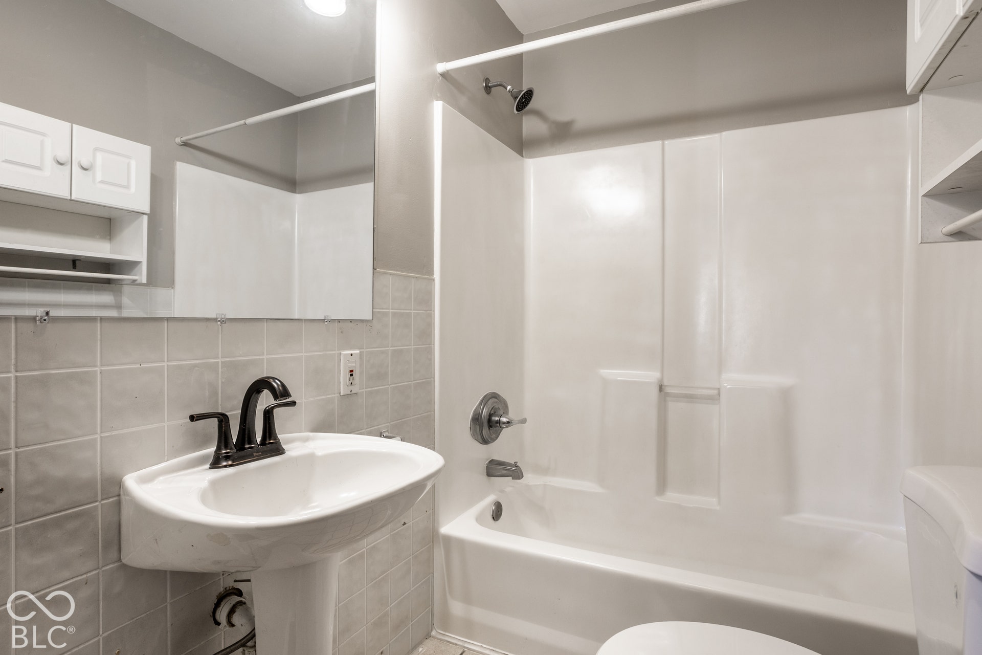 Pedestal sink with a dark bronze-finish faucet, tiled wall, wall-mounted mirror, white wall cabinetry, and a white shower-tub combination unit