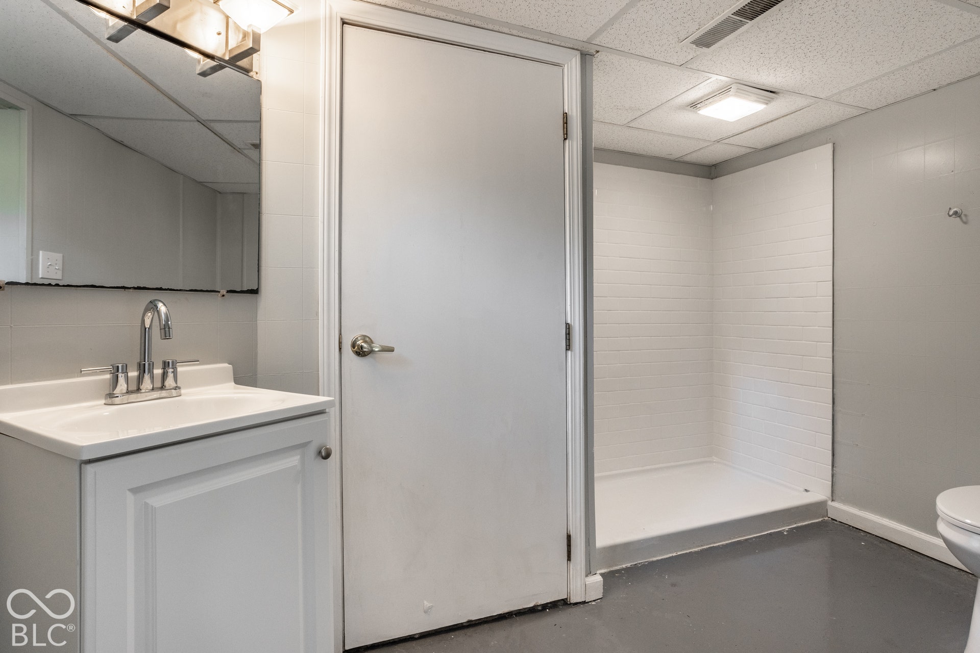 Basement full bathroom - White vanity with integrated sink, chrome faucet, wall-mounted mirror, and overhead vanity lighting