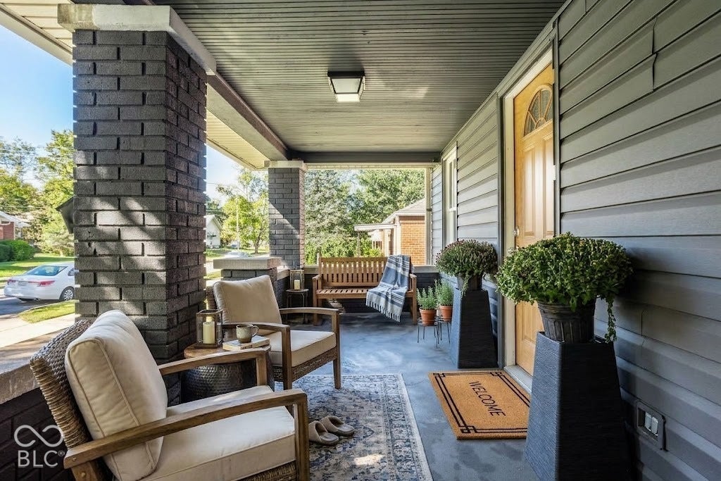 Virtually Altered - Covered front porch featuring dark gray brick pillars, gray horizontal siding, and a wooden entry door with a decorative arch window