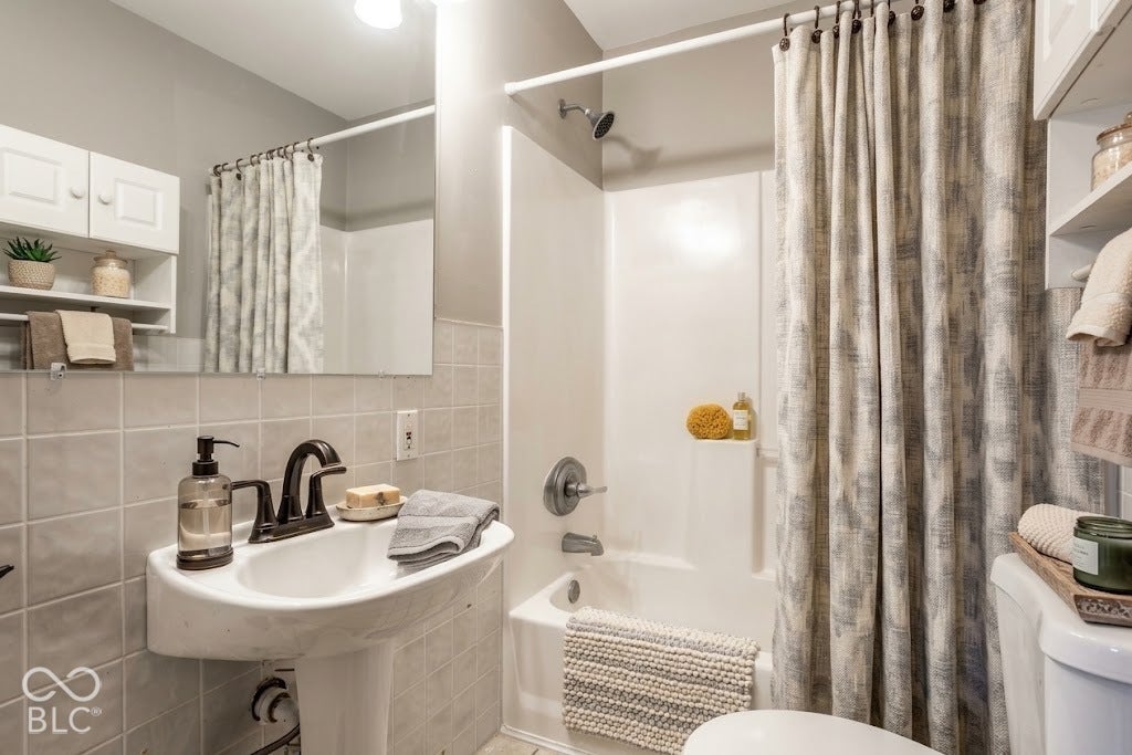 Virtually Altered - Pedestal sink with dark-toned fixtures, tiled walls, built-in tub with shower, and overhead storage cabinetry