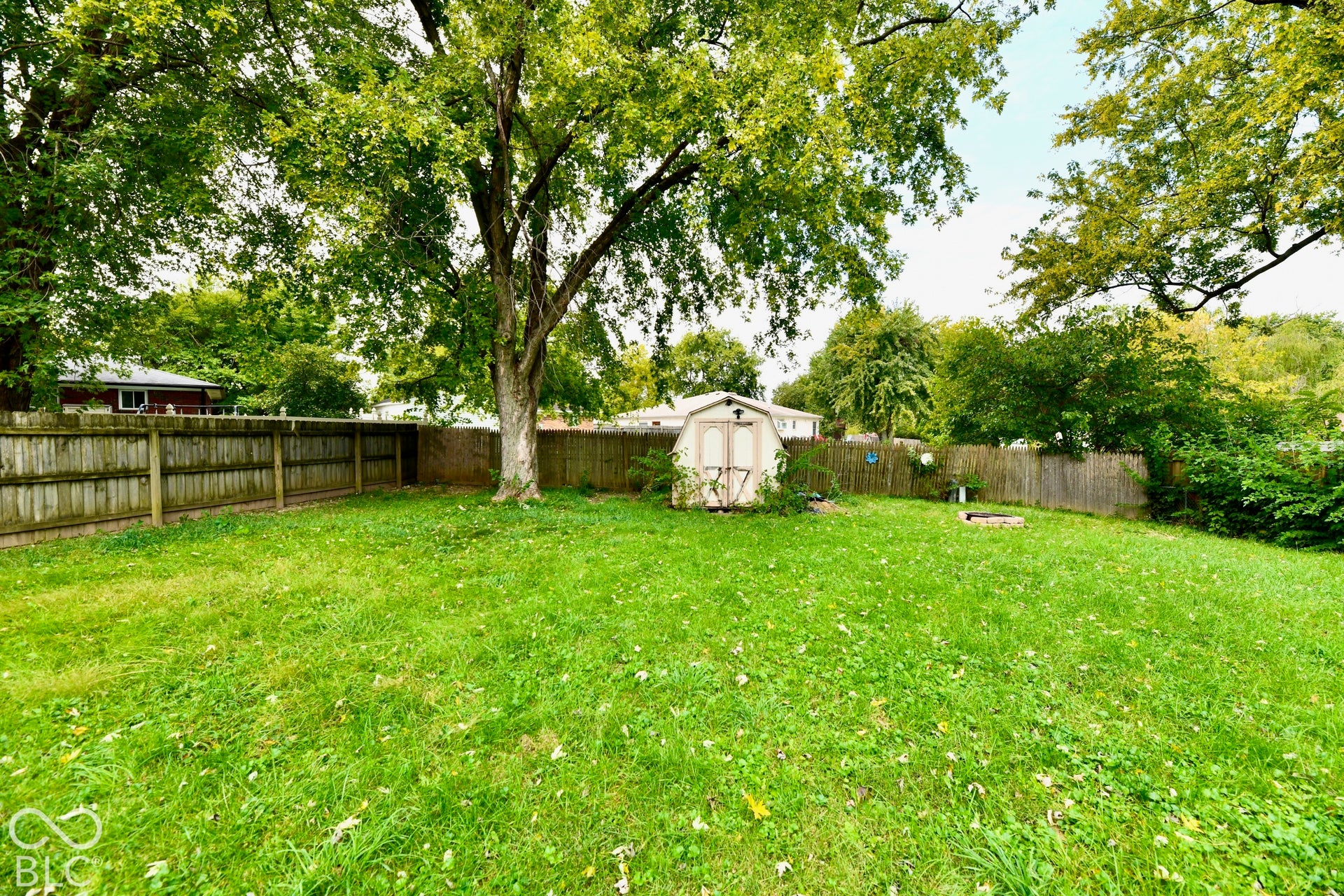Expansive backyard featuring a manicured lawn, established trees, wood fencing, a storage shed, and a fire pit