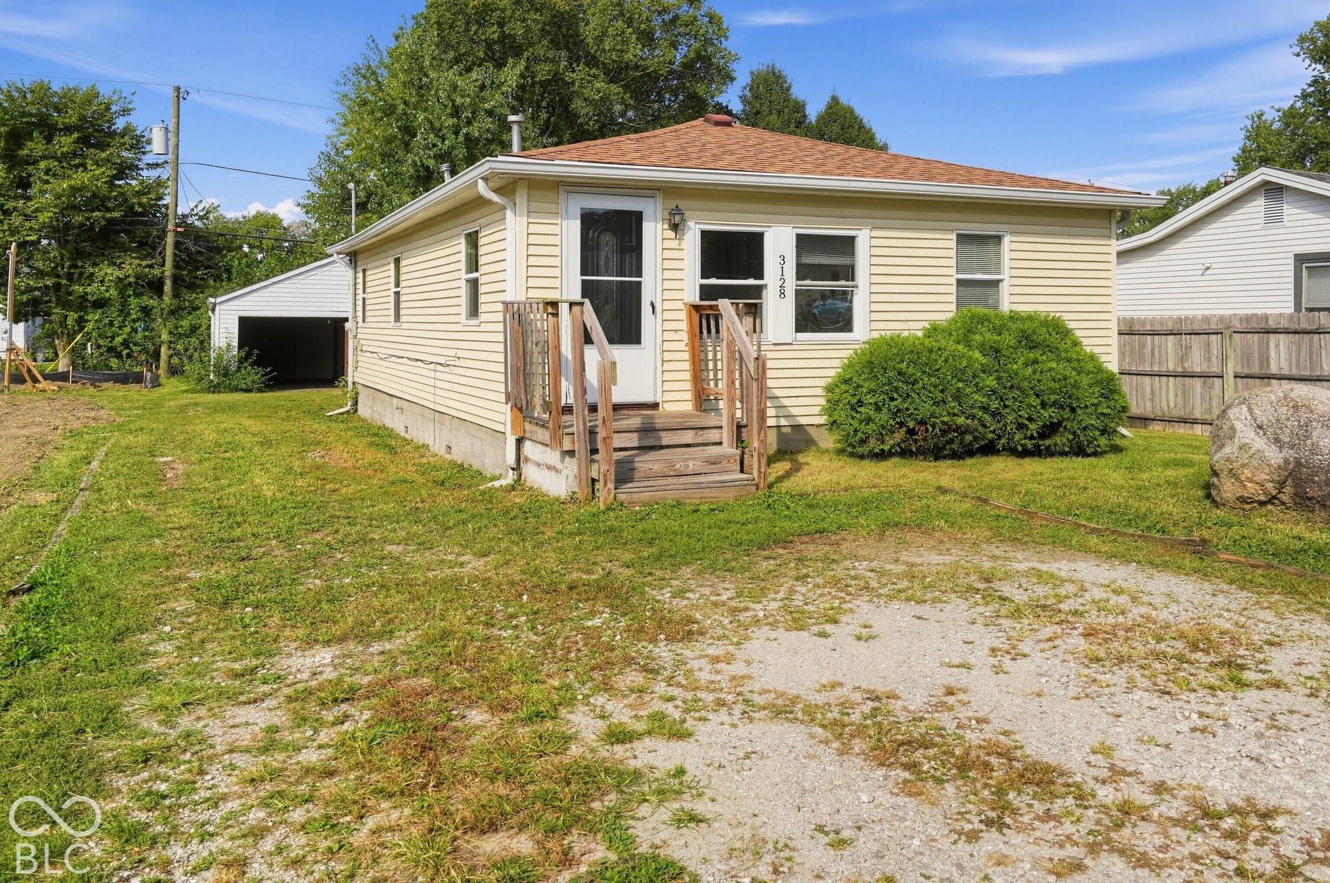 3128 S Rural Street, Indianapolis, IN 46237 Light-colored siding exterior, wood deck, gravel parking area, separate detached garage structure, and established shrubbery