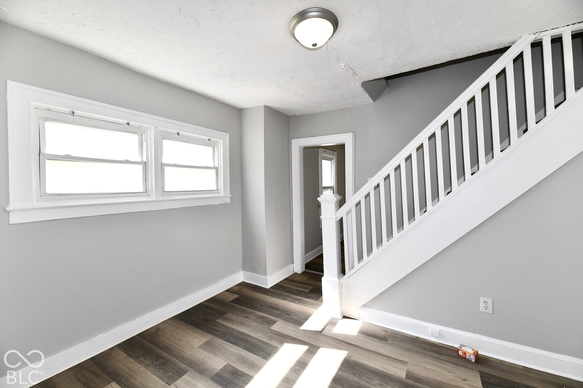 Wood-finish flooring with a light gray wall color, two windows with white trim, and a white staircase with balusters