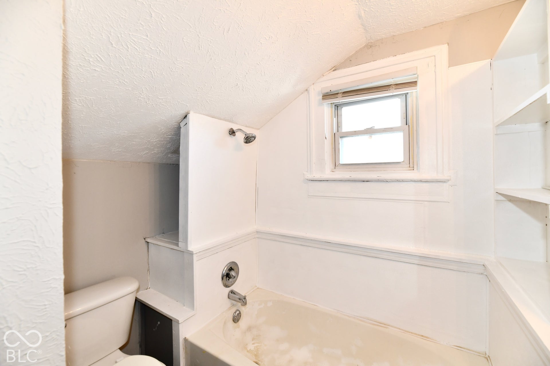 Built-in bathtub with shower fixture, window with blinds, and integrated shelving