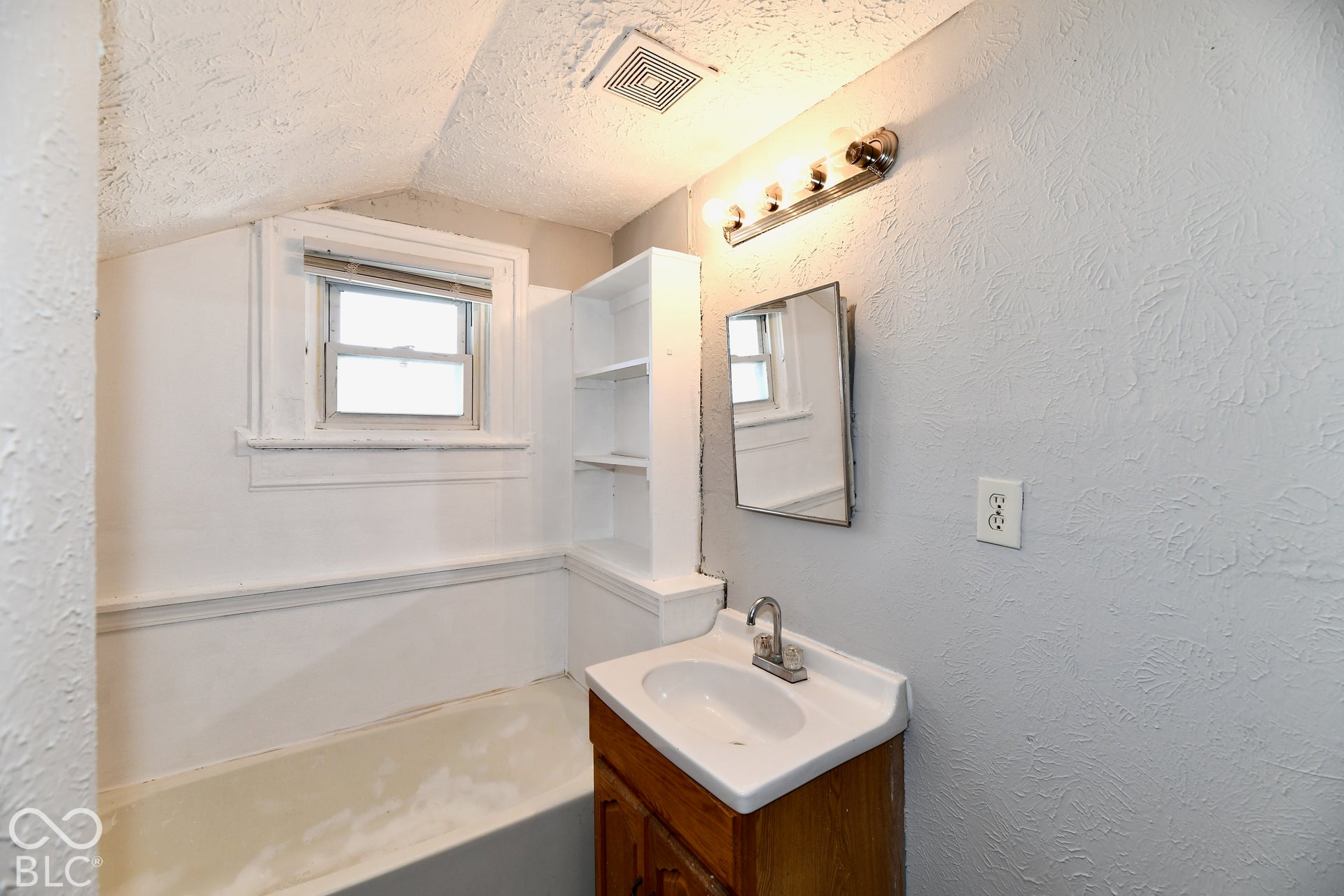 White paneled tub with window, built-in shelving, and a vanity with a basin sink
